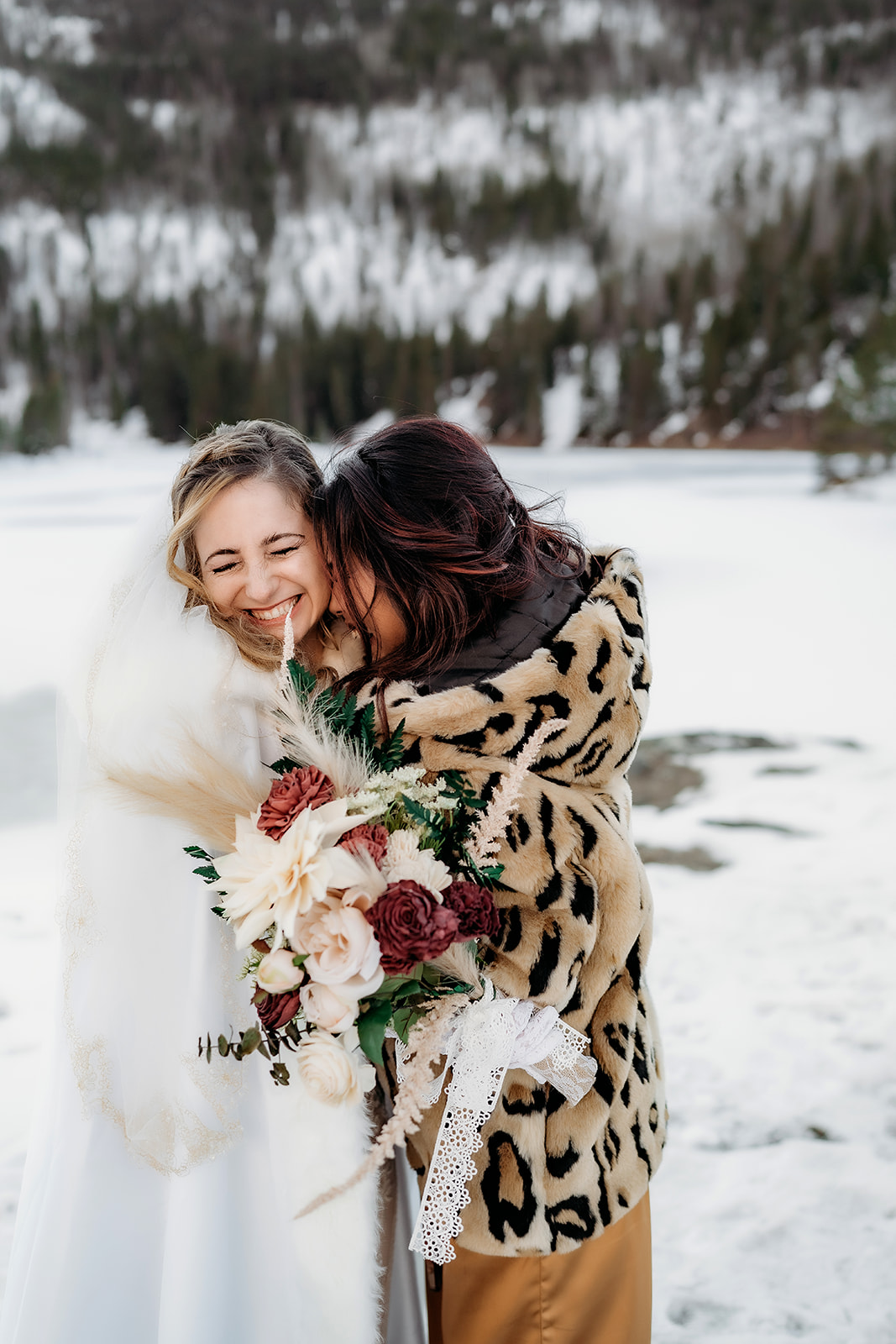 Bride laughs in a warm embrace with a guest wearing a leopard coat, holding her winter floral bouquet.