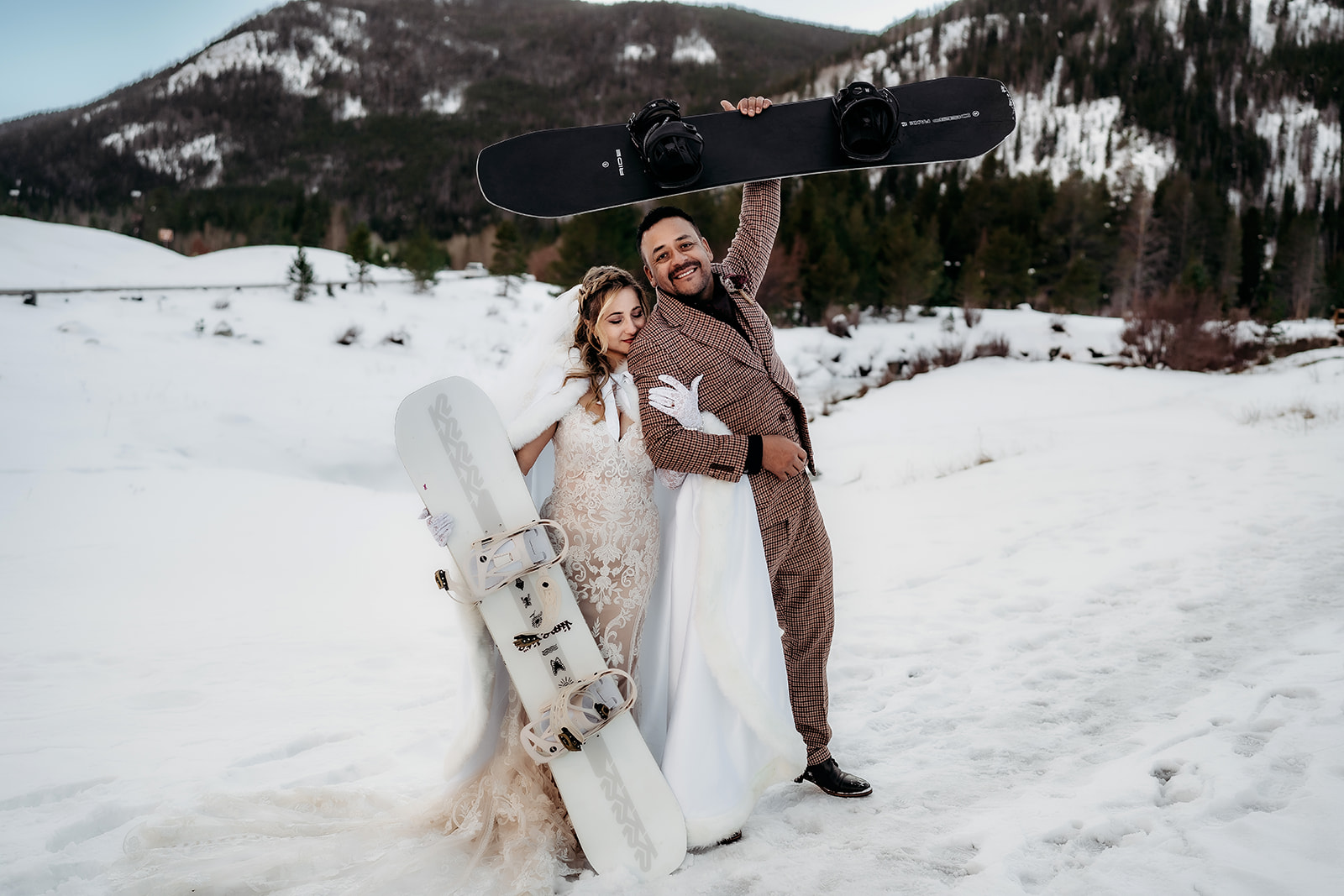 Bride and groom pose playfully with snowboards in hand, celebrating their adventurous winter elopement in Colorado with snow-covered mountains behind them.