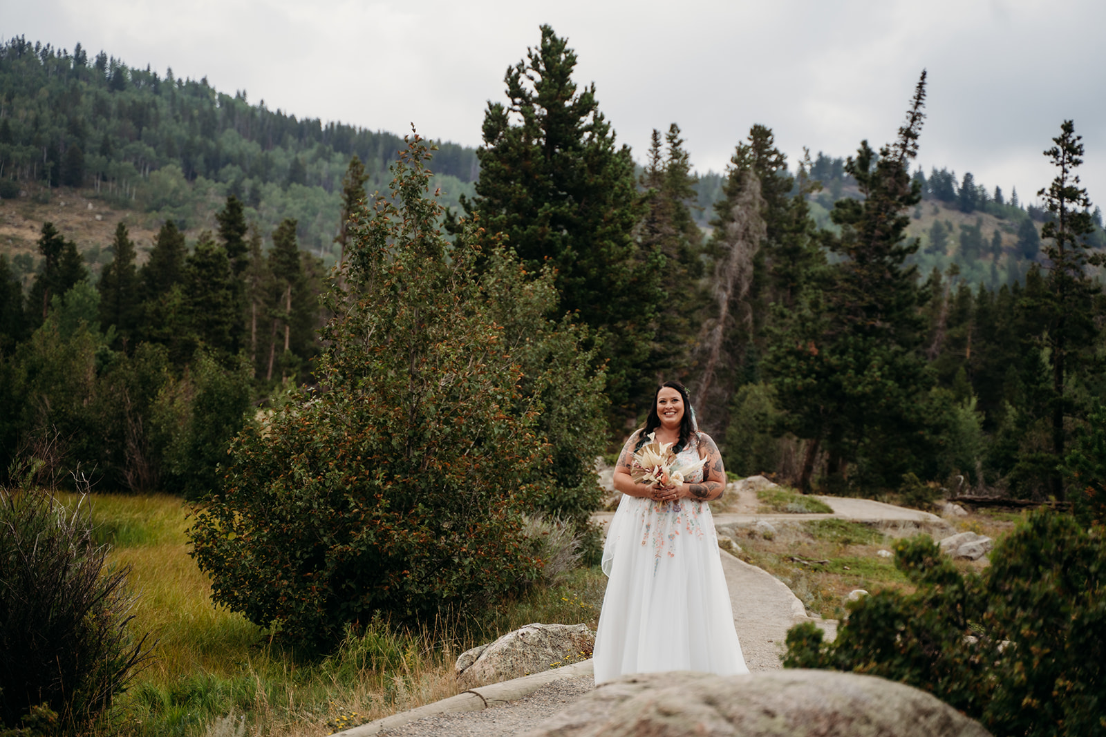 Bride smiles while standing alone on a winding path through the mountains, holding her bouquet before the ceremony.