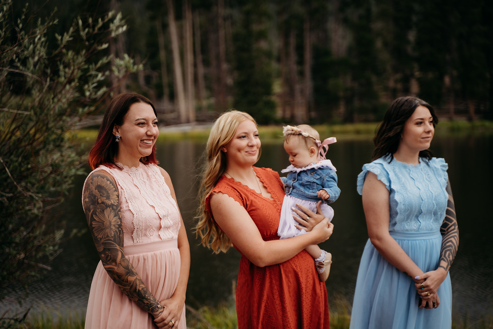 Three women and a baby stand near a quiet lakeshore in soft pastel and earthy-toned dresses, smiling before the ceremony.