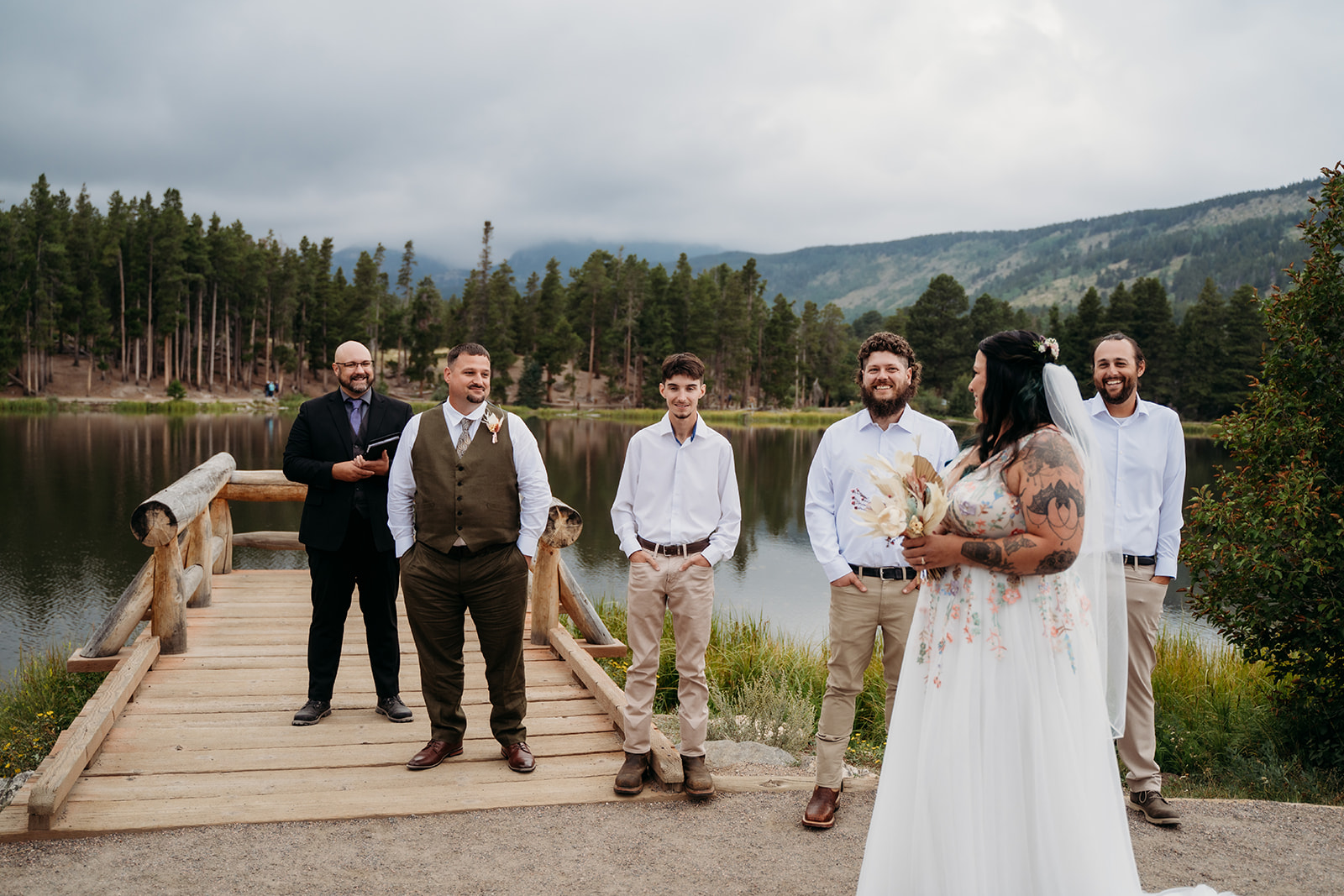 Bride approaches her groom and their small wedding party on a rustic dock by a quiet lake, surrounded by forested mountains.