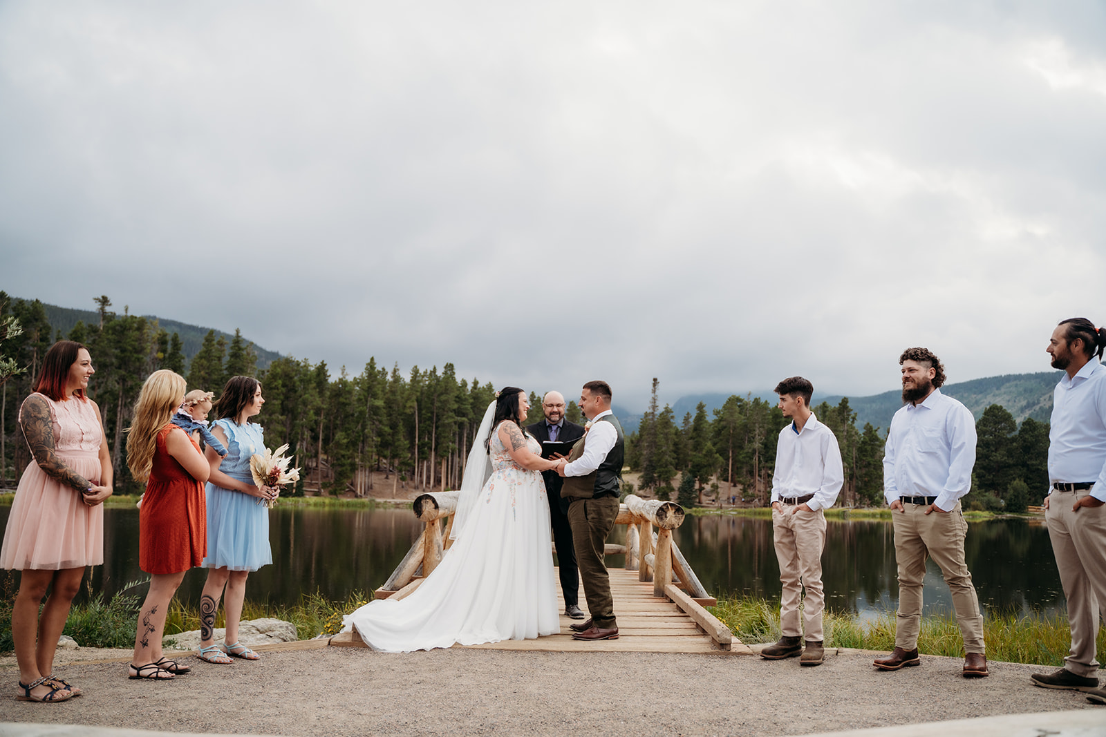 Couple exchanges vows on a dock, surrounded by loved ones and pine trees during their intimate wedding ceremony.