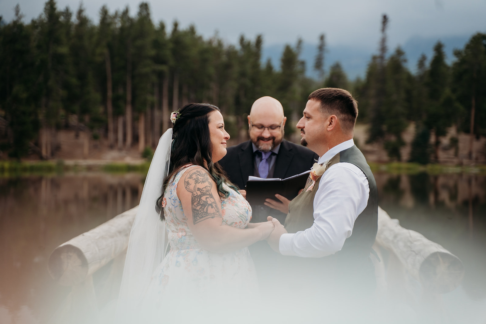 Close-up of the bride and groom holding hands and exchanging vows on a wooden dock surrounded by water and trees.