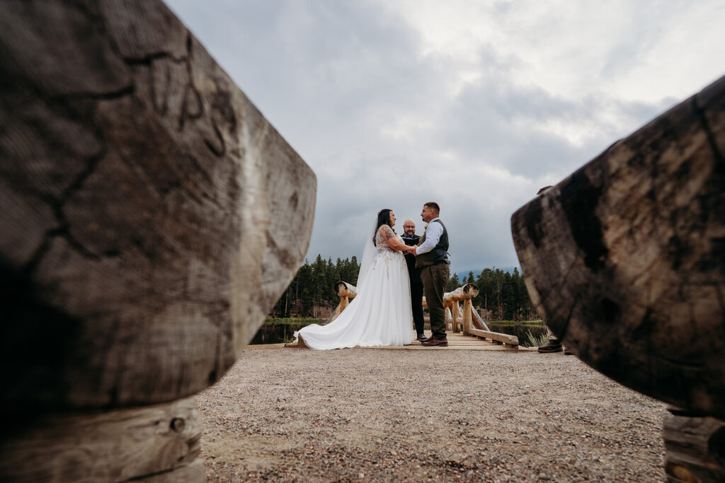 Wide-angle view of the couple standing on the dock, holding hands during their intimate wedding ceremony with cloudy skies above.