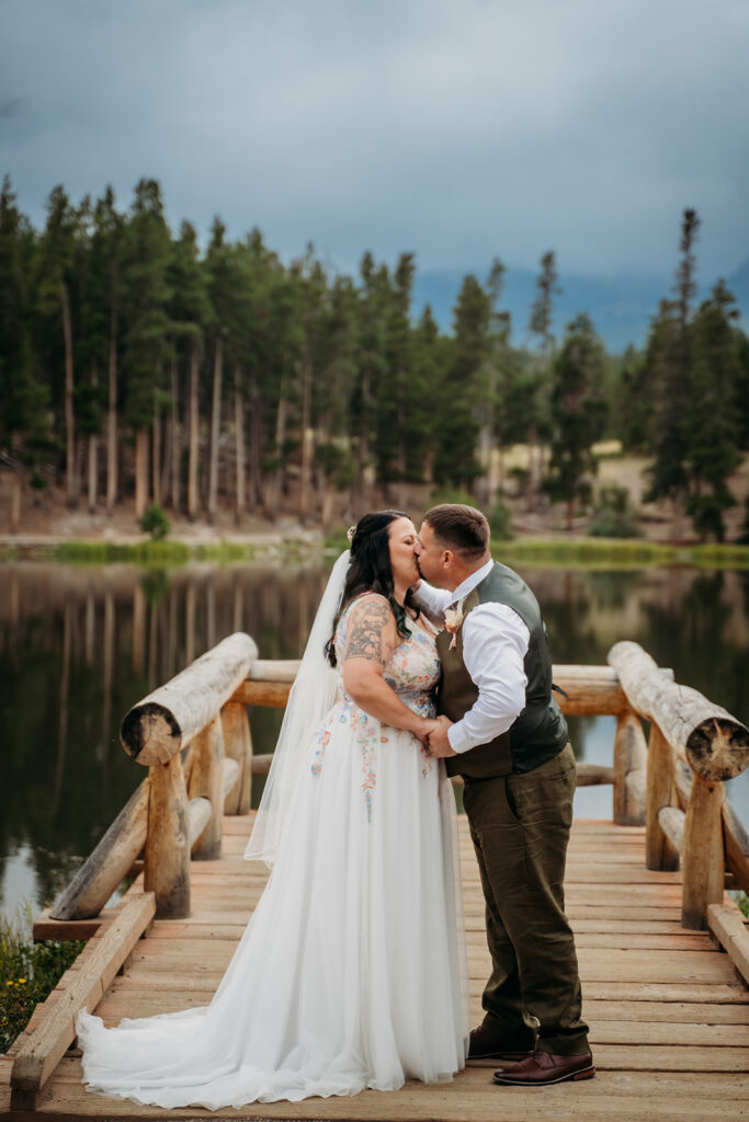 Bride and groom share their first kiss as a married couple, standing on a rustic wooden dock at the edge of the lake.