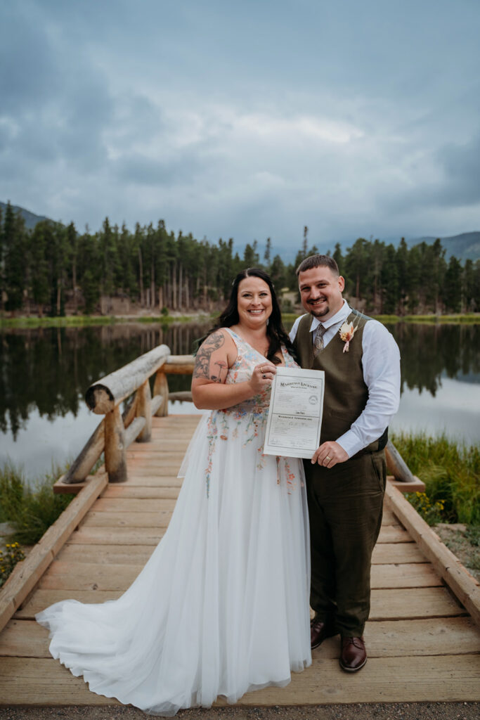 Bride and groom proudly hold their signed marriage license on the dock with pine trees and still water behind them.