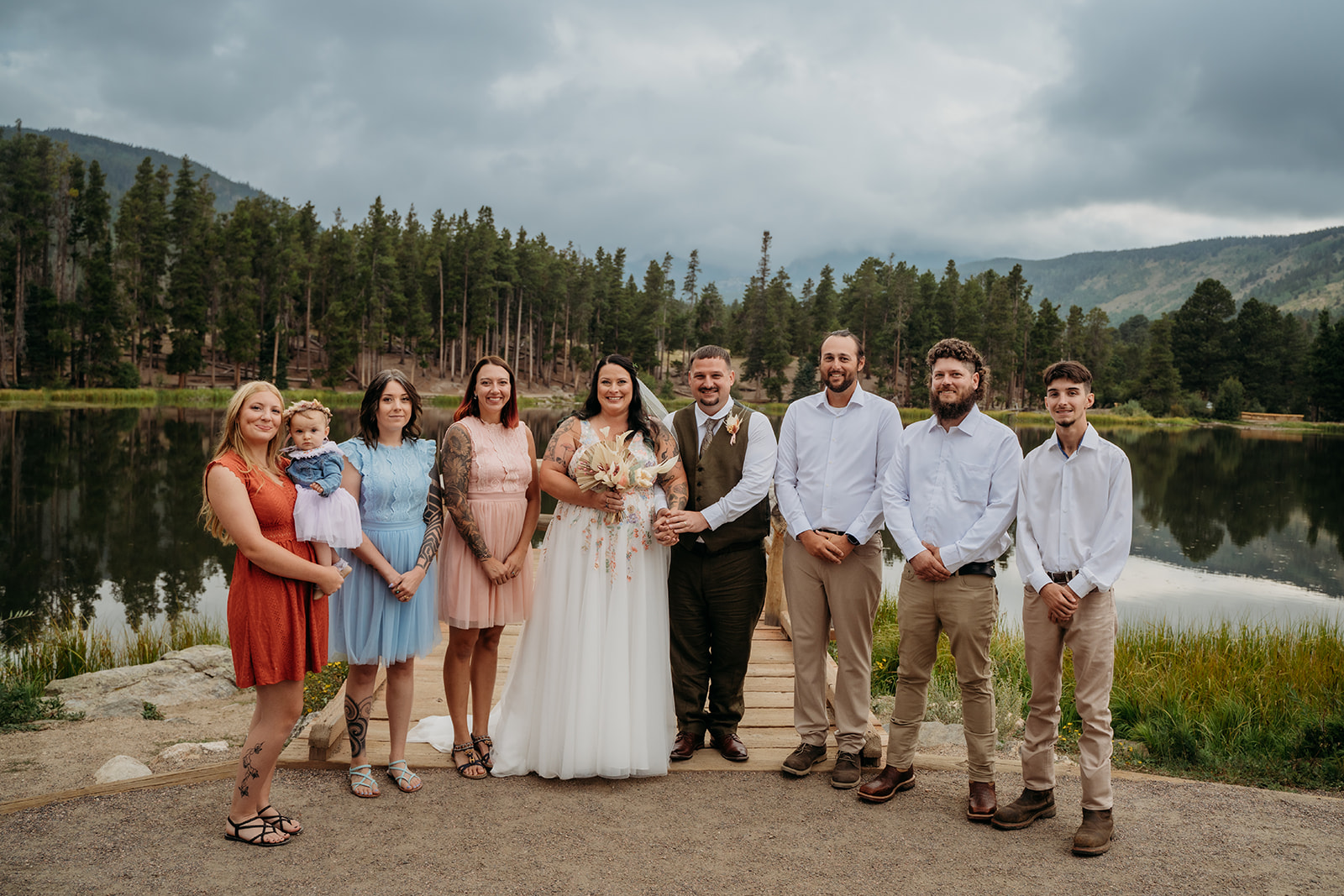 Bride and groom pose with their wedding party in front of a serene mountain lake, surrounded by loved ones and cloudy skies.