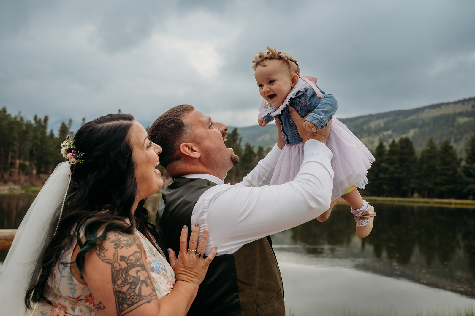 Groom lifts baby girl into the air while the bride smiles up at her, capturing a joyful moment from their intimate wedding.