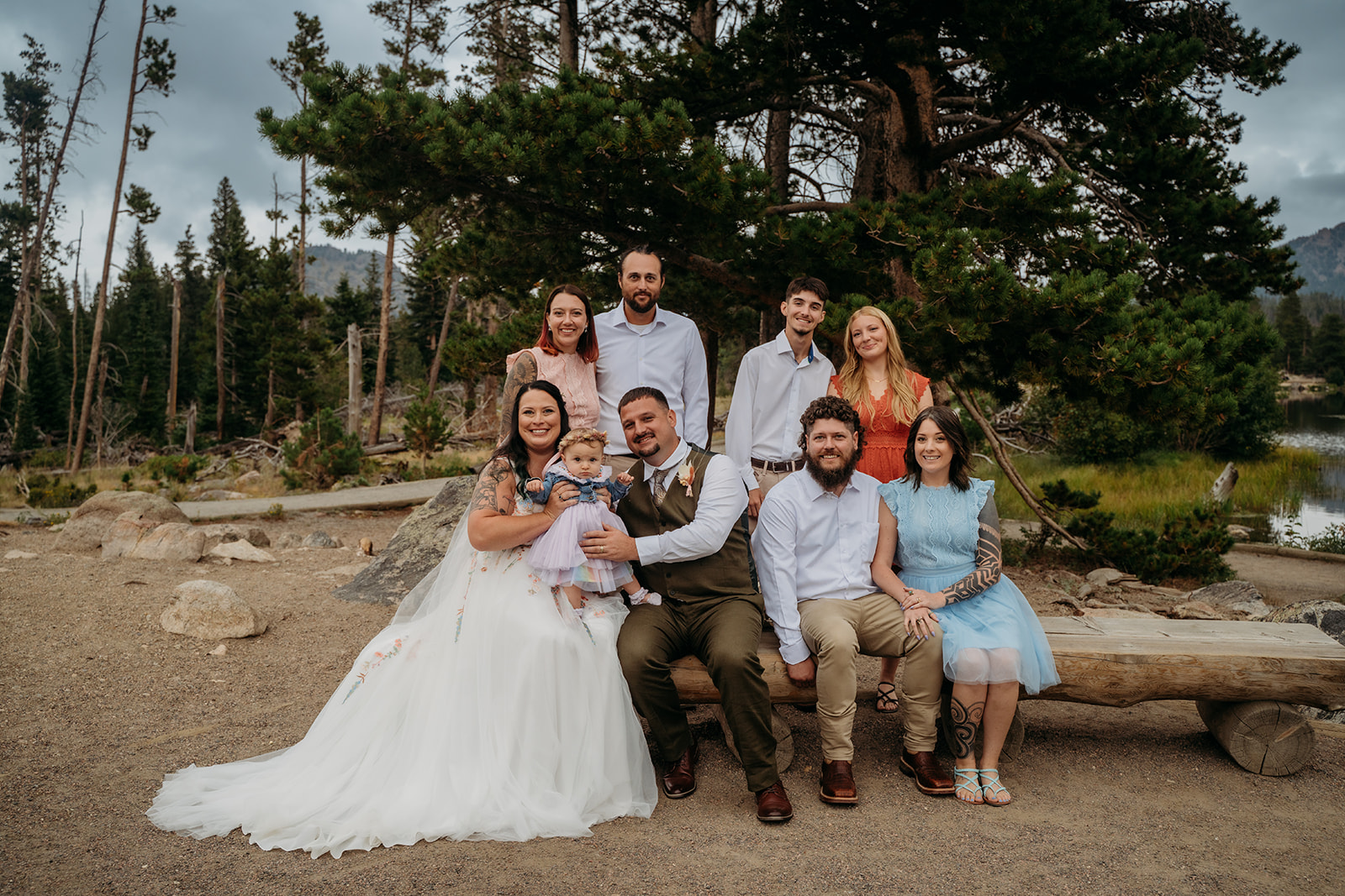 Full family portrait of the wedding couple and their guests, gathered together outdoors beneath tall pines.