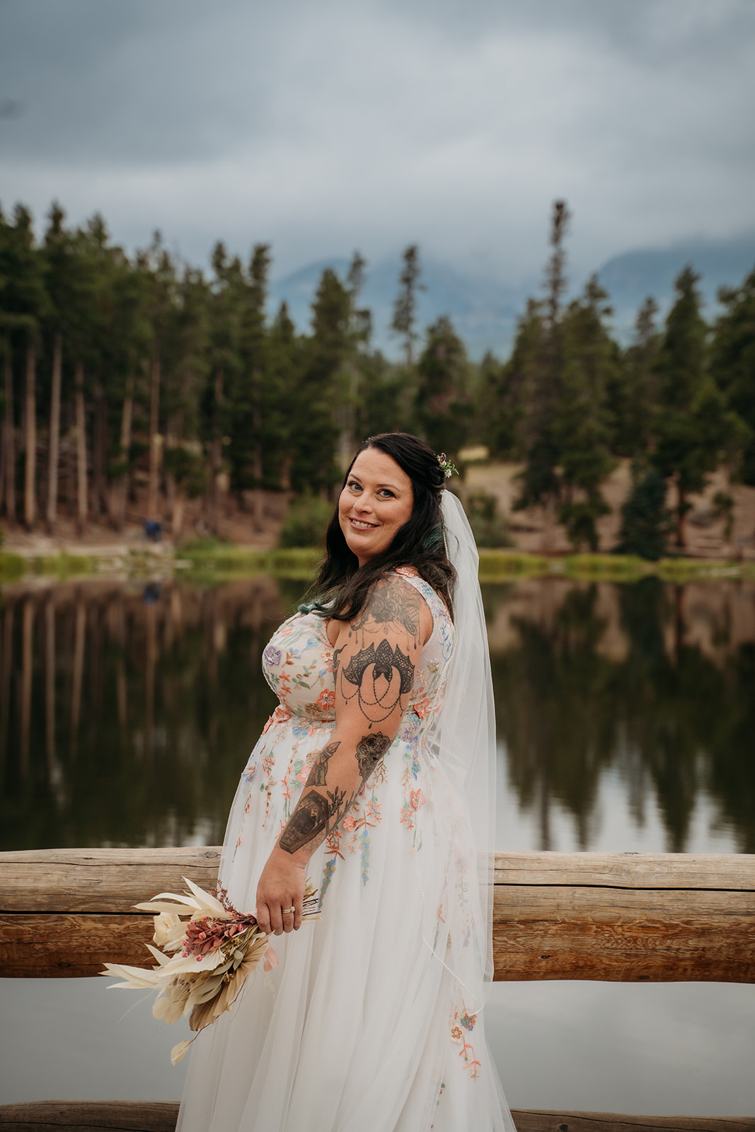 Bride stands smiling by the lake with her bouquet in hand, framed by trees and the soft reflection of mountain peaks.
