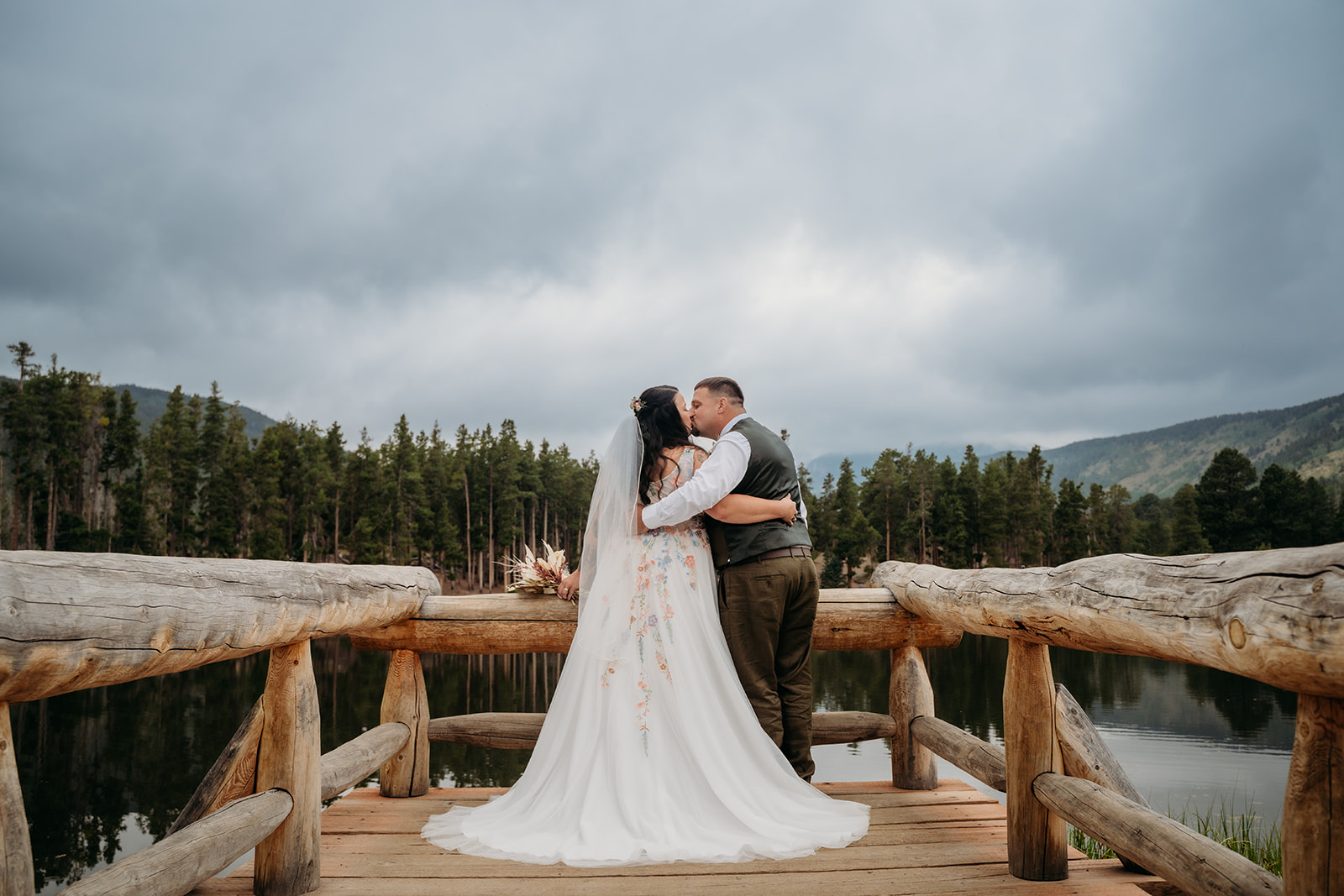 Bride and groom kiss at the edge of the dock, surrounded by evergreens and dramatic skies on their lakeside intimate wedding day.