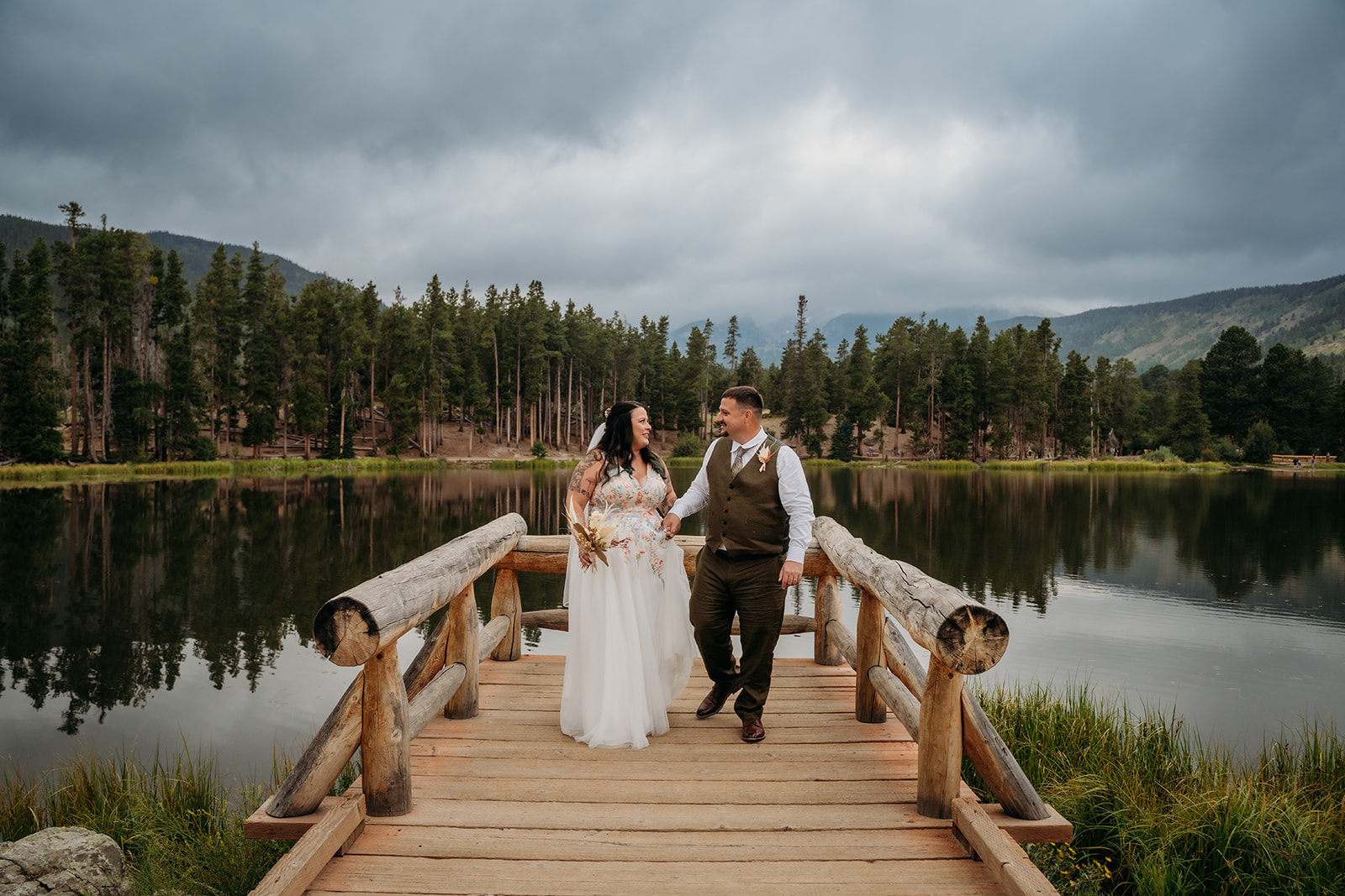 Bride and groom walk hand-in-hand across a wooden bridge over a lake, smiling during their intimate wedding celebration.