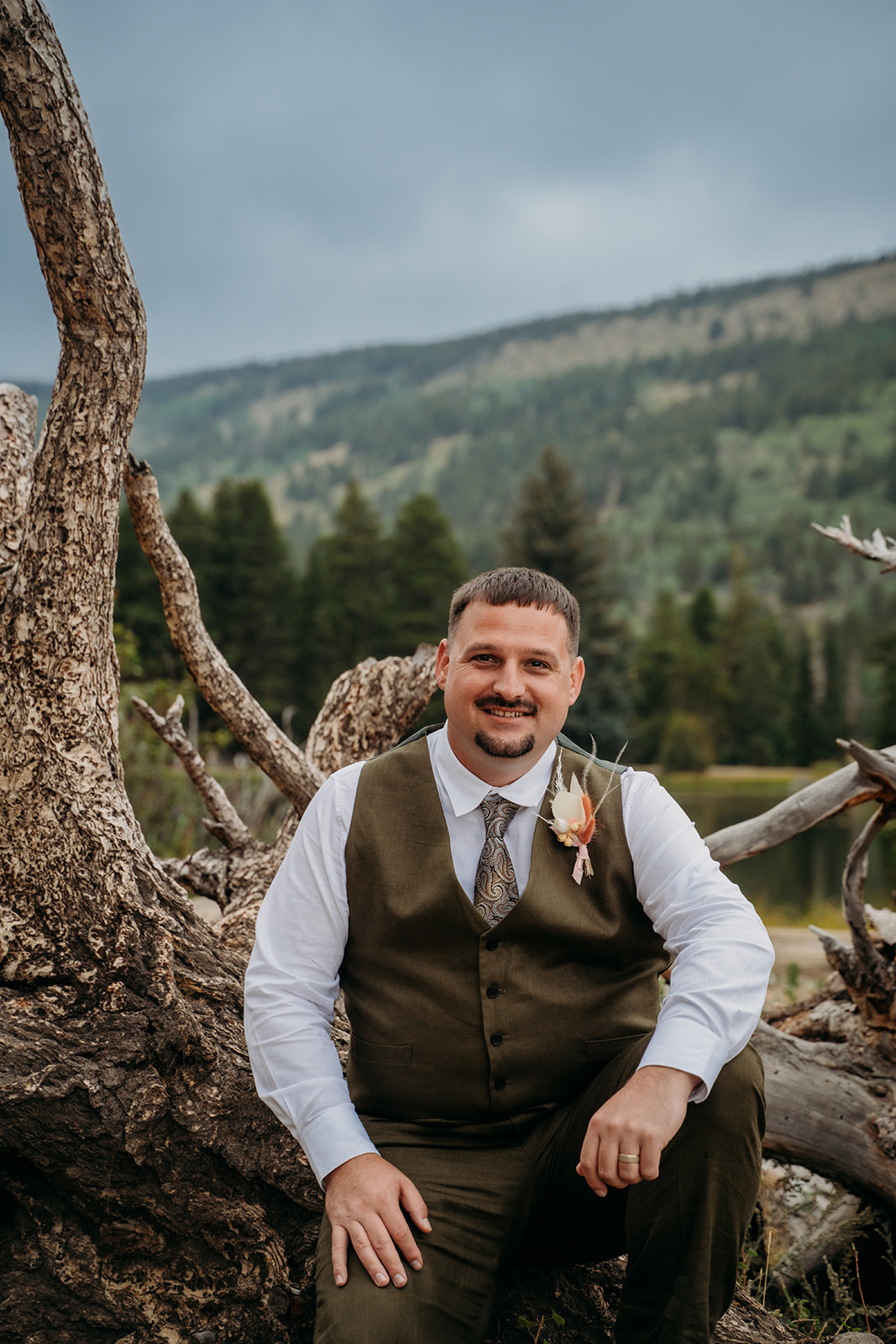 Groom sits beside a twisted tree trunk, smiling at the camera with forested hills behind him.
