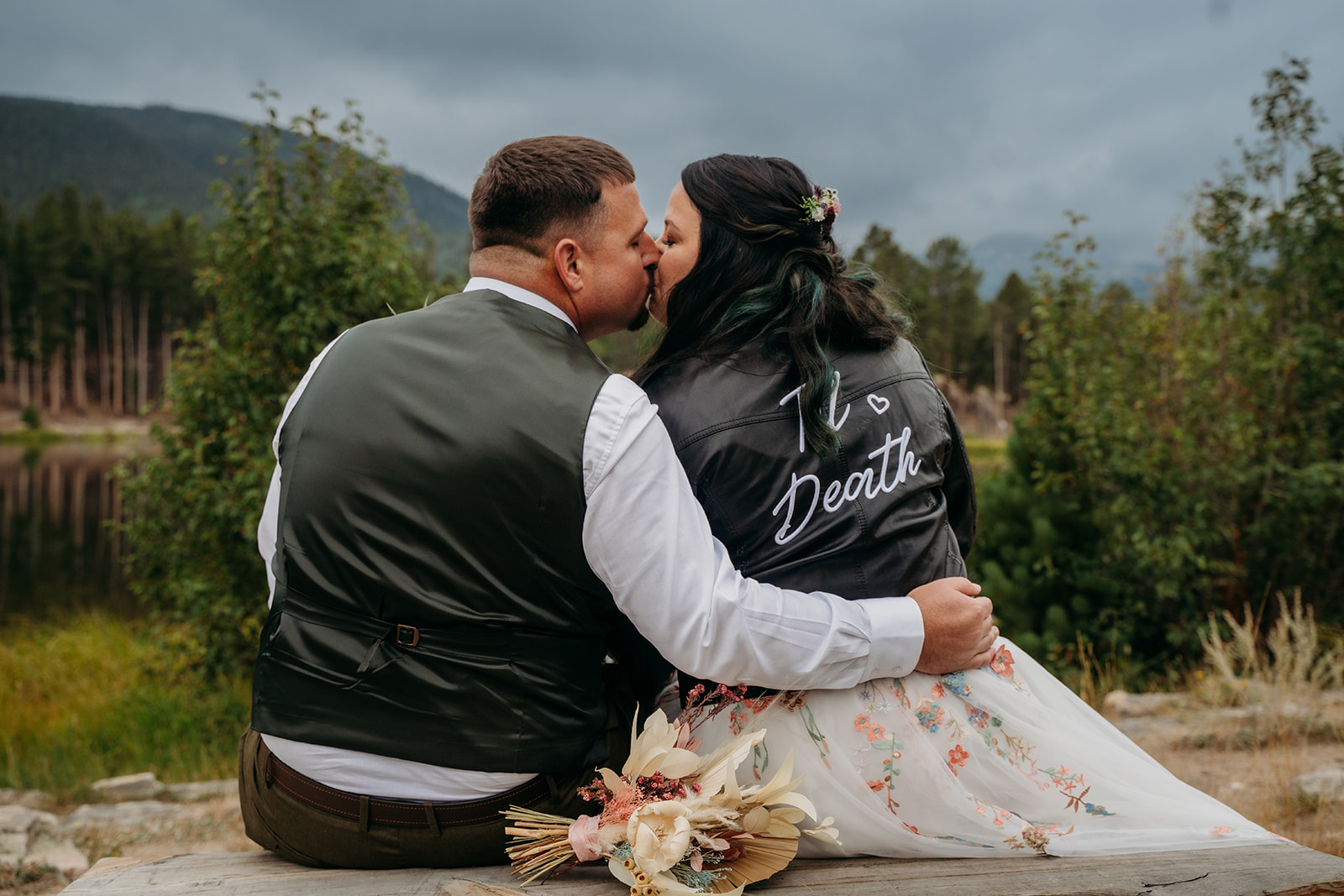 Bride and groom kiss while seated, both wearing matching "Til Death" jackets with the bouquet placed nearby.