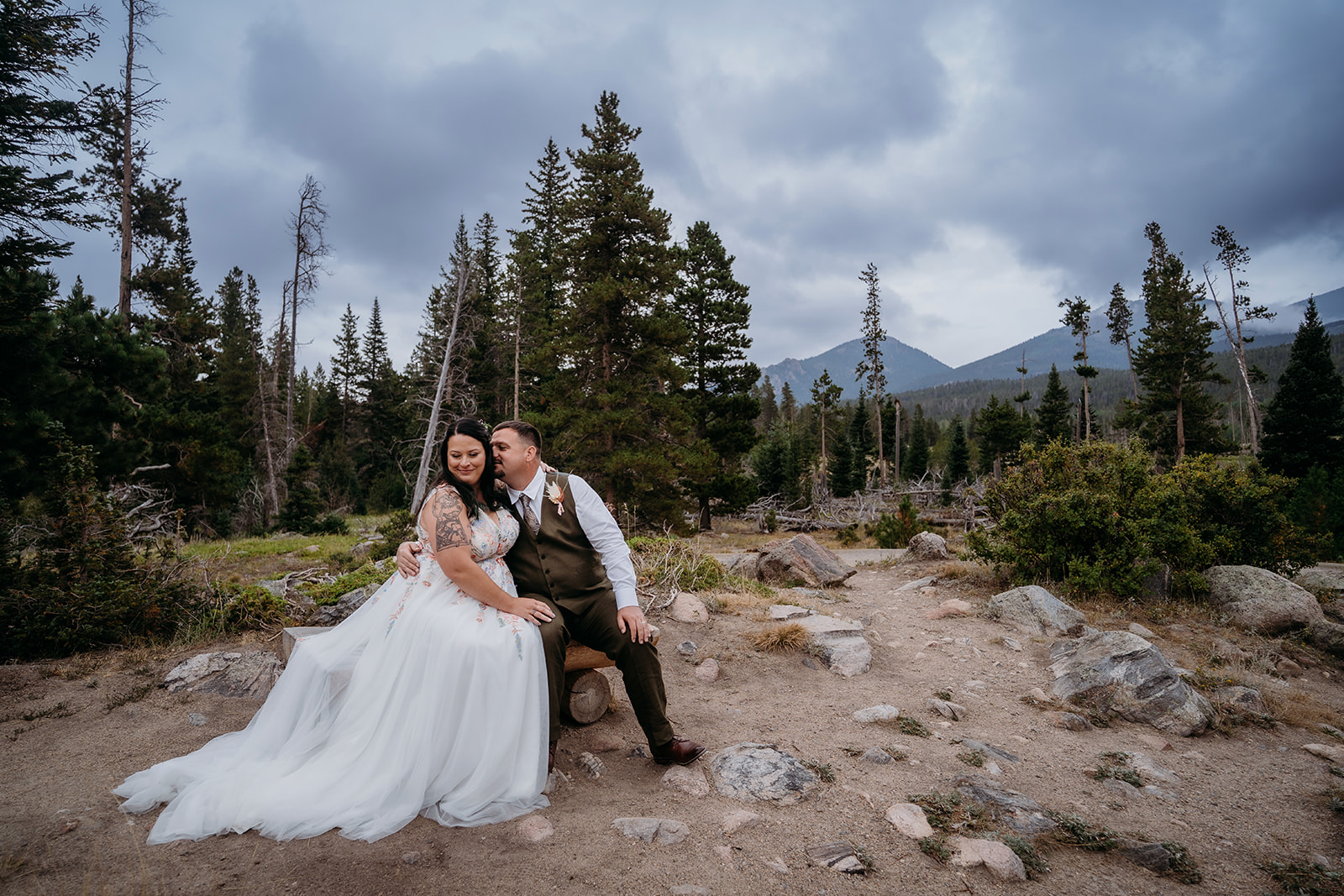 Bride sits on groom’s lap as they share a quiet moment in a rocky mountain clearing surrounded by tall pines.