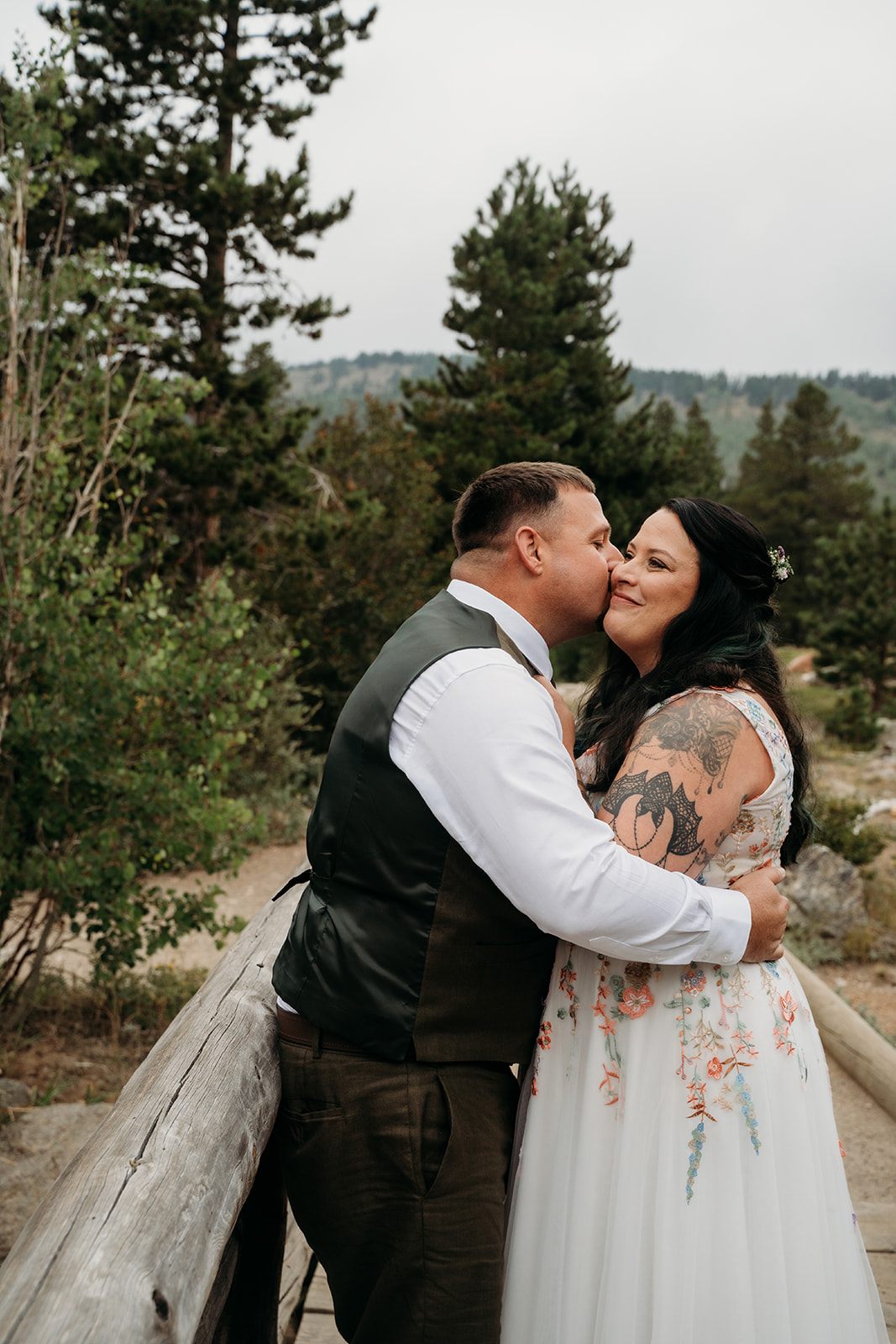 Groom kisses the bride’s cheek as they embrace beside a rustic wooden fence with alpine trees in the background
