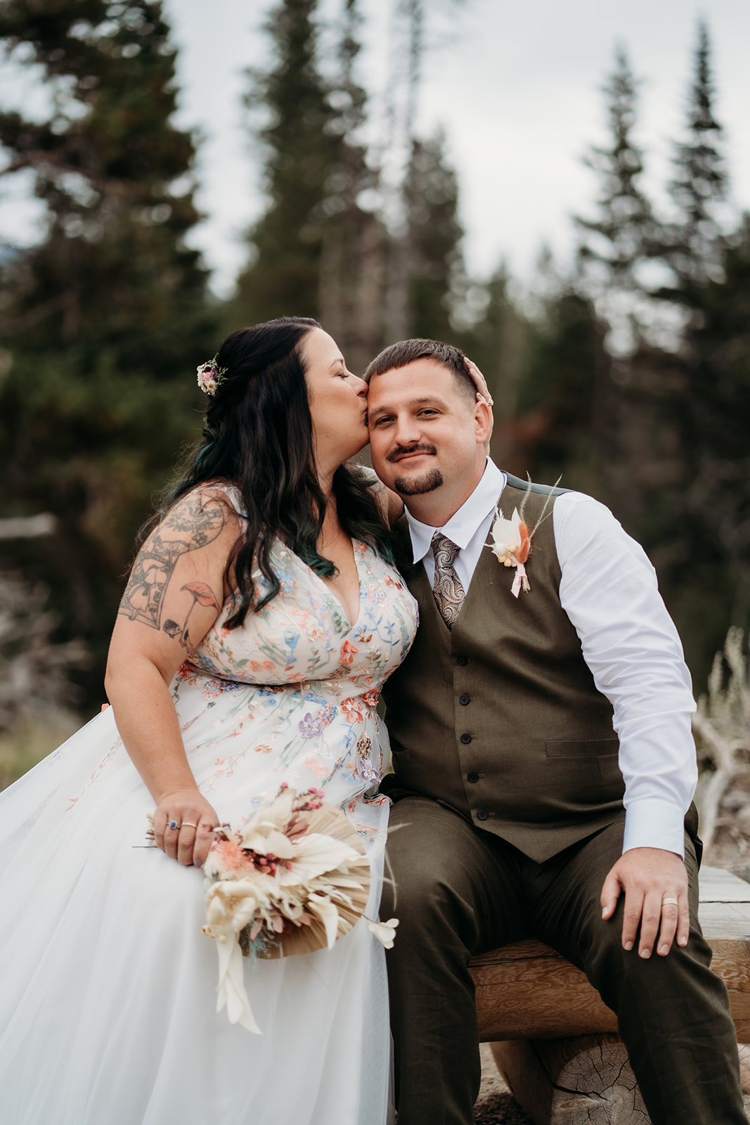 Bride kisses the groom on the temple as they sit close together on a rustic bench during their lakeside wedding celebration.