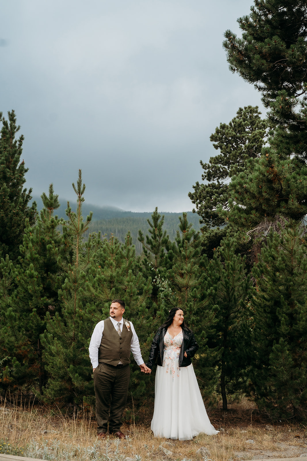 Bride in a leather jacket and groom holding hands in a pine forest, embracing the moody mountain vibes of their wedding day.