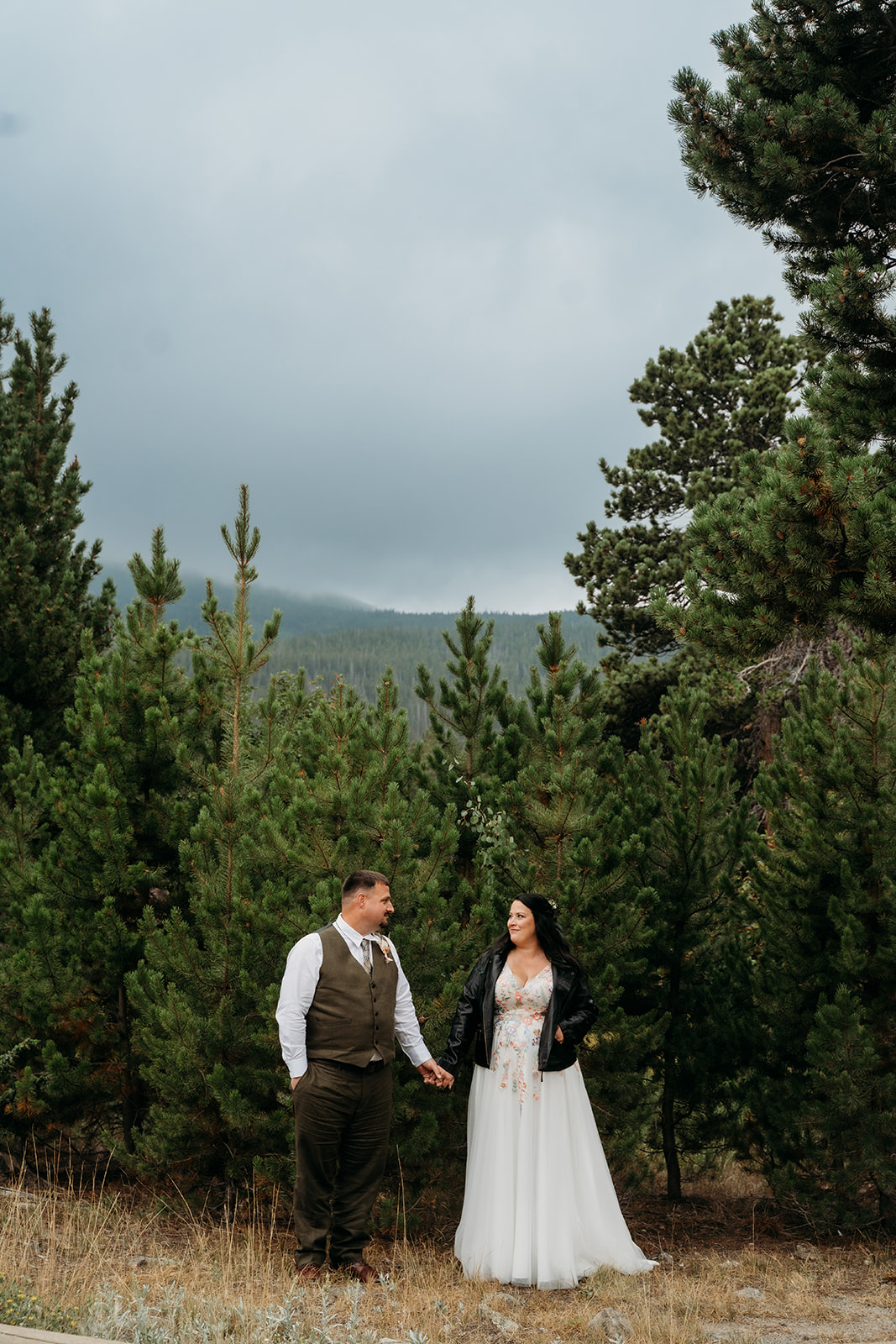 Bride and groom hold hands in a pine forest, her leather jacket adding a cozy touch to their adventurous Colorado elopement.