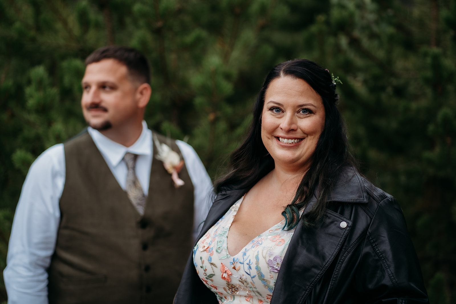 Bride stands in the foreground smiling confidently while the groom gazes off behind her, both dressed in unique wedding attire.