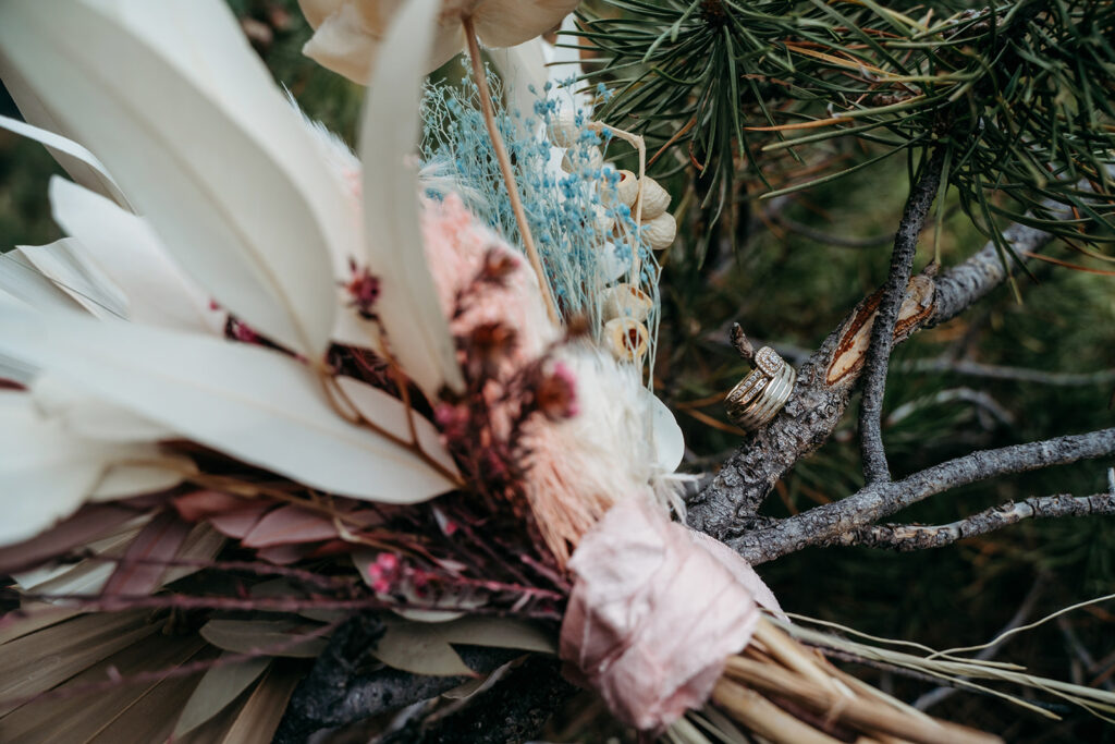 Close-up of the bride’s bouquet with dried florals and soft pastels, resting beside tree branches and wedding rings.