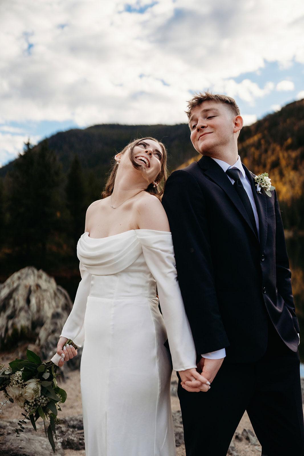 The bride throws her head back in laughter as she and her groom pose side by side in front of an alpine lake.