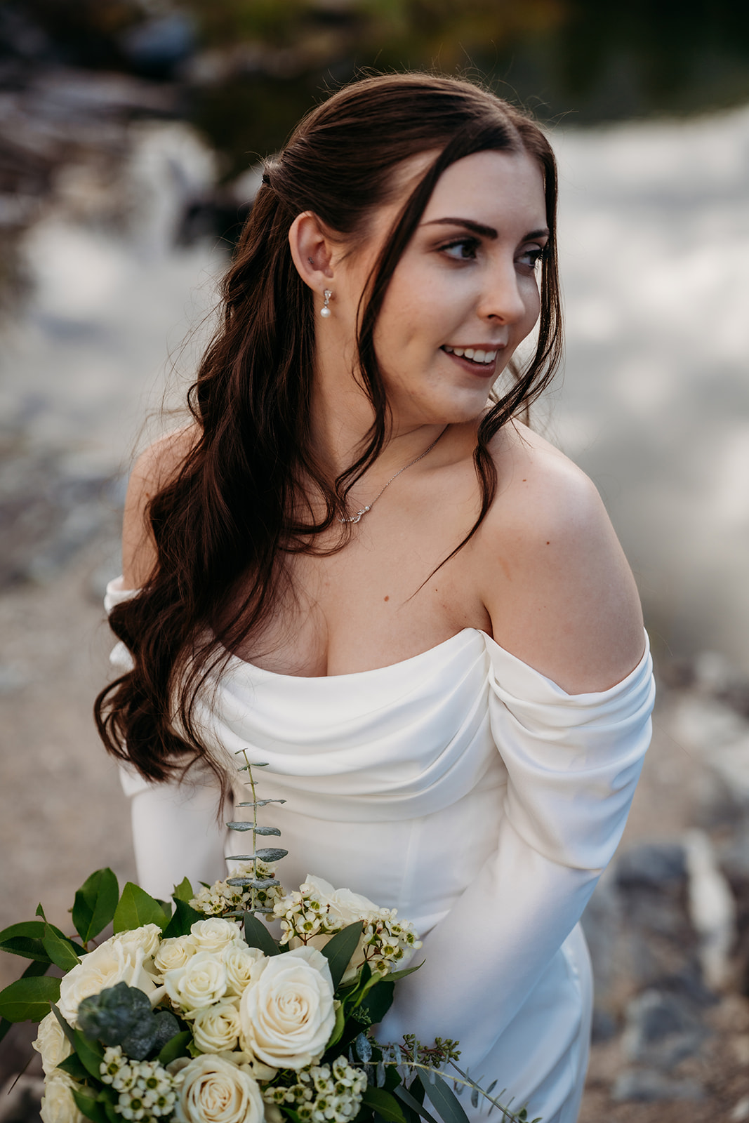 The bride glances off to the side, bouquet in hand, with the soft river and mountain light highlighting her natural beauty.