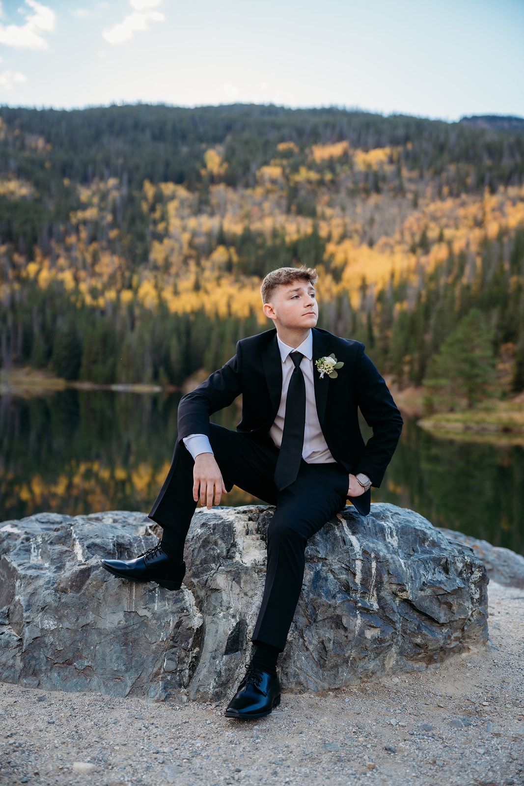 The groom sits on a boulder with fall colors behind him, pausing for a portrait before the ceremony.