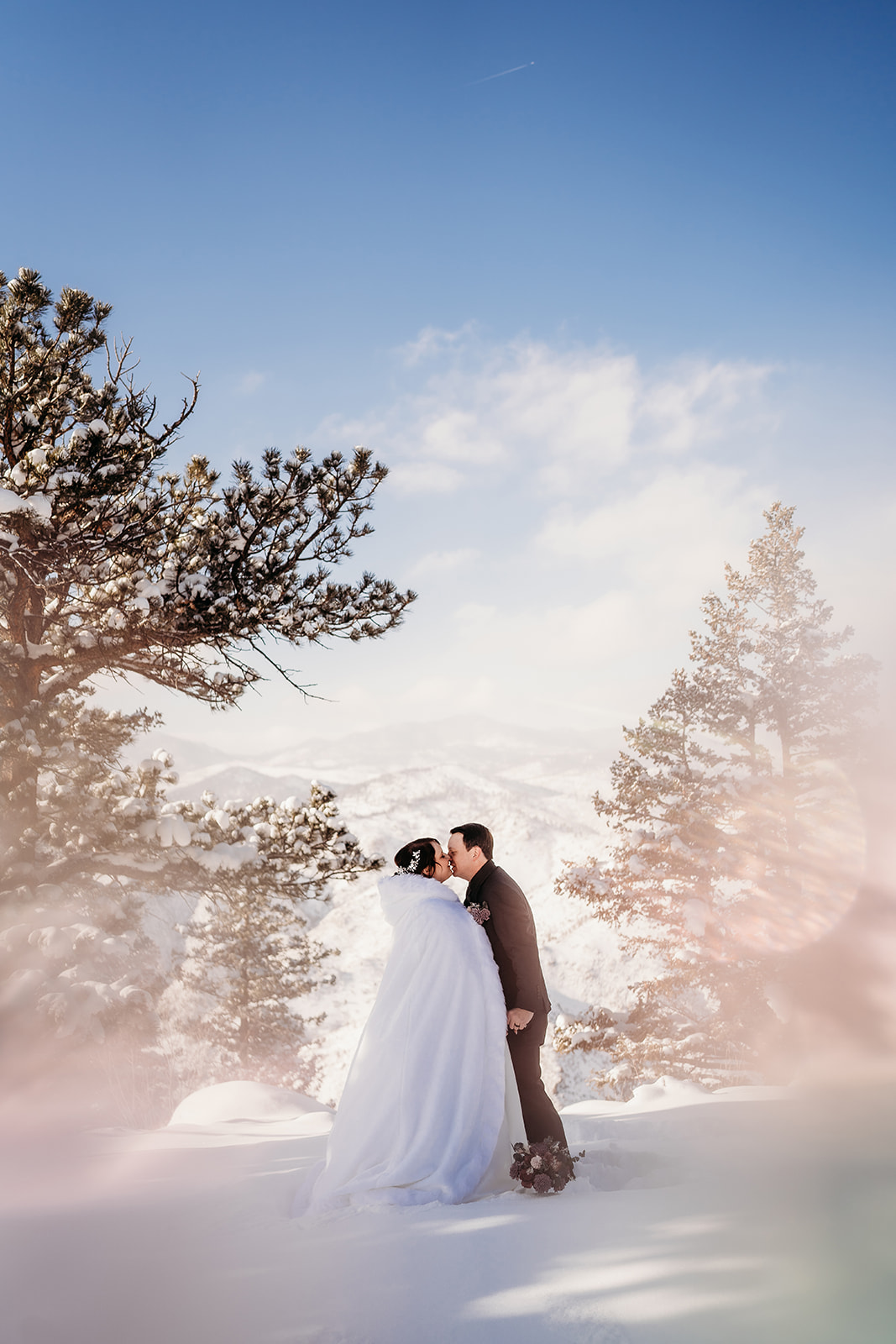 Bride in a full-skirted gown and groom in a black suit sharing a quiet moment at sunset with snow-covered mountains behind them.