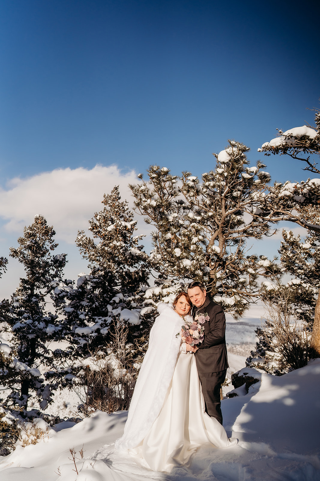 Smiling couple wrapped in a winter cape and suit, framed by snow-covered trees under a bright blue Colorado sky.