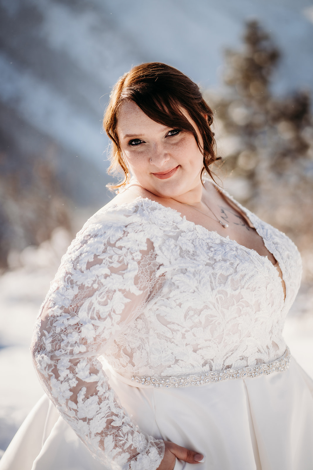Bride in a long-sleeve lace wedding dress smiling softly while standing in the snow with a mountain backdrop.