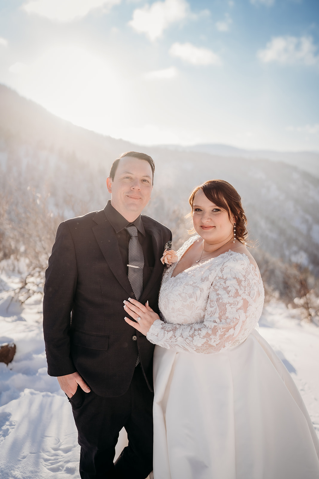 Couple smiling at the camera, the bride in a long-sleeved lace dress and the groom in a black suit surrounded by glowing winter light.