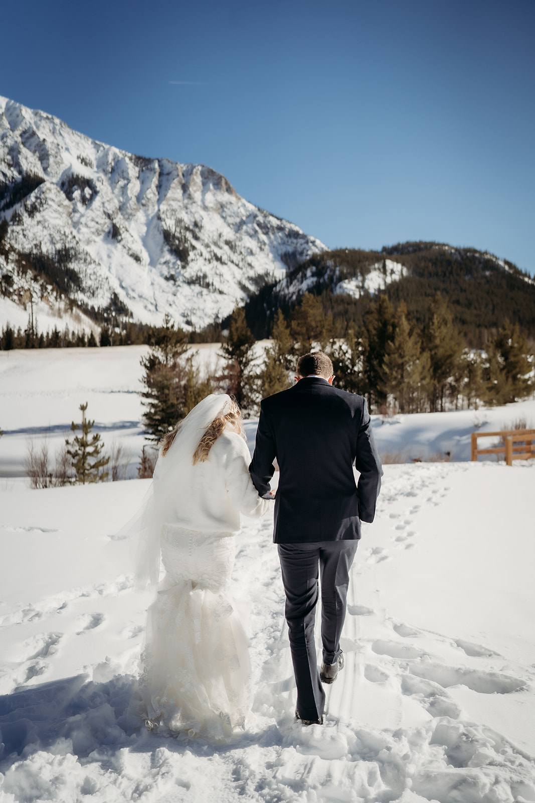 Bride and groom walk toward snow-covered Colorado peaks, the bride’s veil and fitted gown adding to their elegant winter wedding outfits.