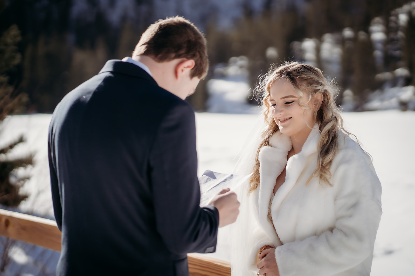 Bride in a white faux fur jacket smiling as the groom reads vows, a touching elopement moment in the Colorado snow.
