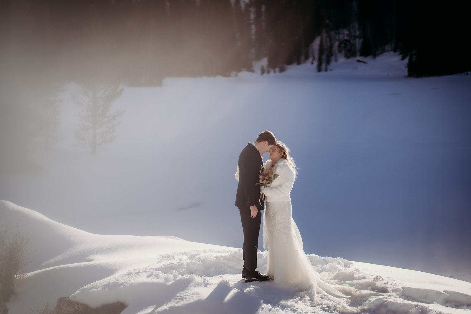 Bride in a long lace gown and faux fur jacket embraces her groom on a snowy ledge—an intimate display of romantic winter wedding outfits.