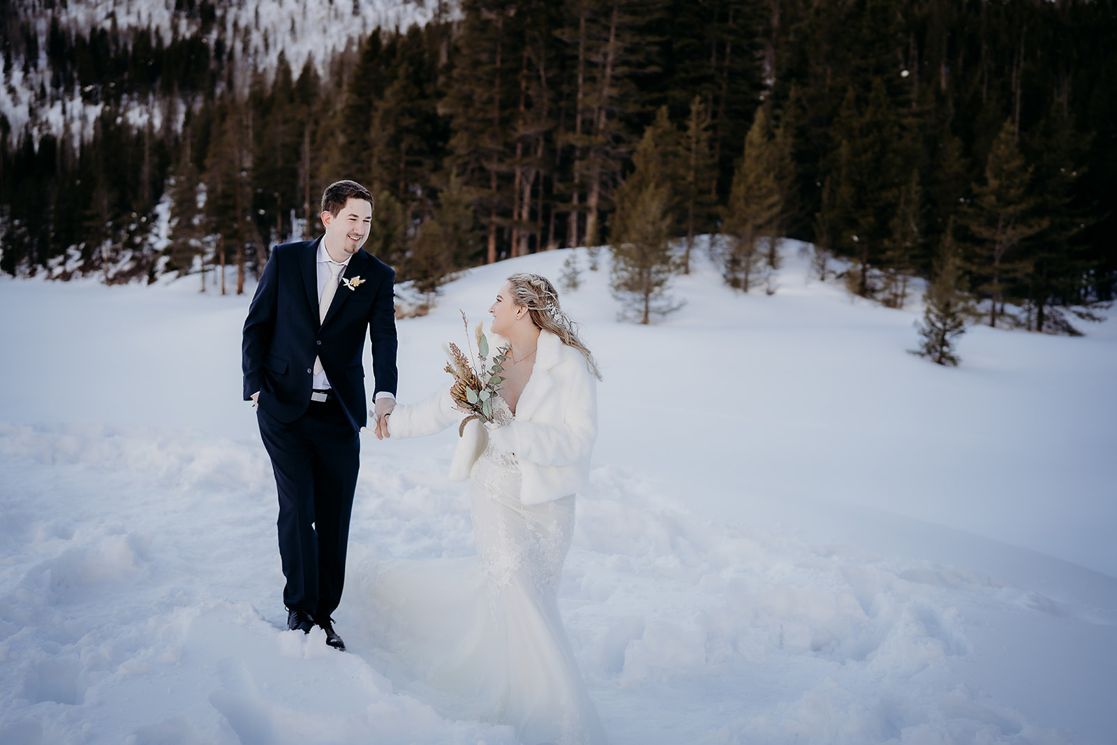 Joyful bride and groom holding hands in fresh snow, with the bride wearing a white faux fur jacket—one of many beautiful winter wedding outfit ideas.