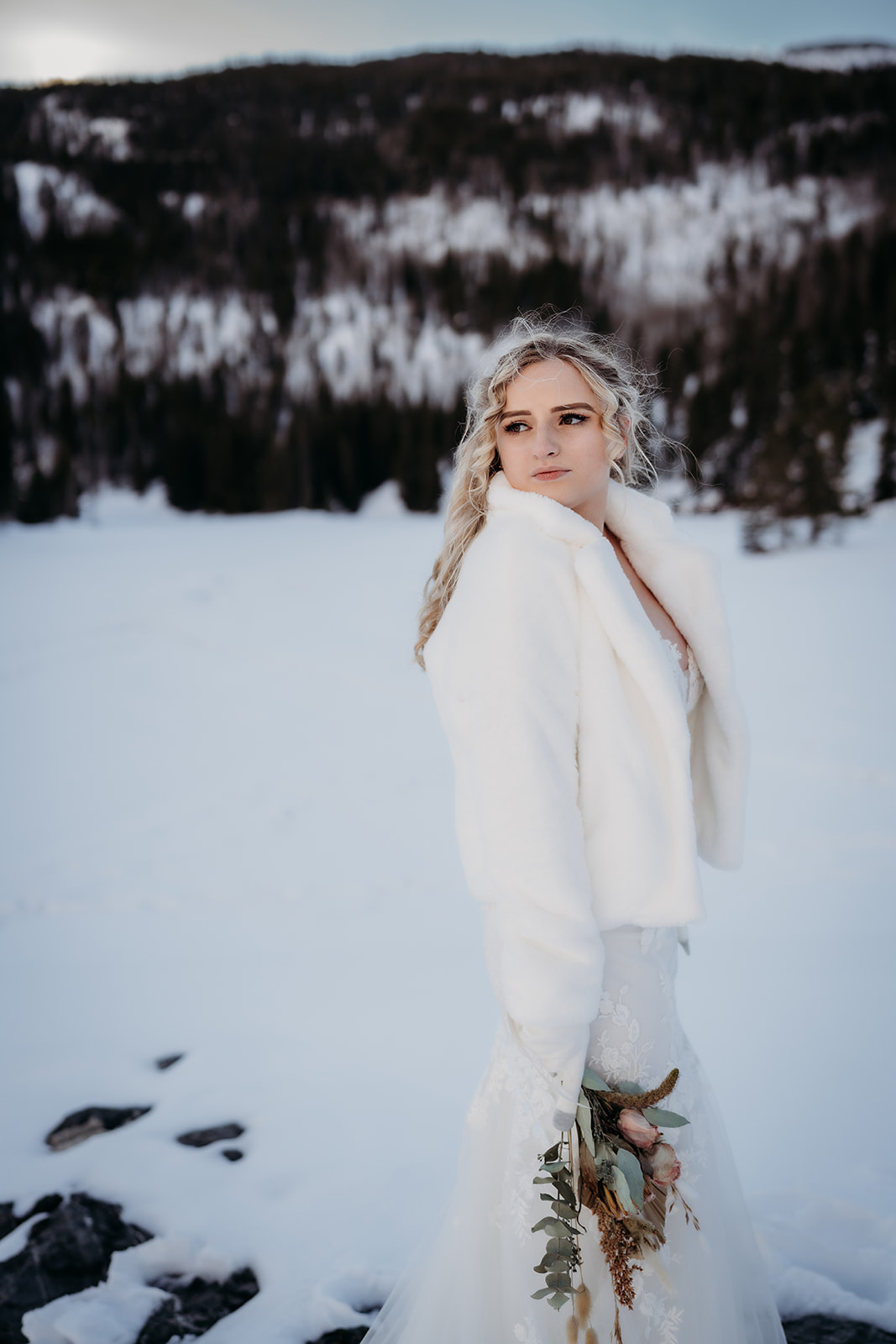 Bride holding a rustic bouquet while standing in a white faux fur coat, the snowy mountain scene highlighting her cozy winter wedding outfit.