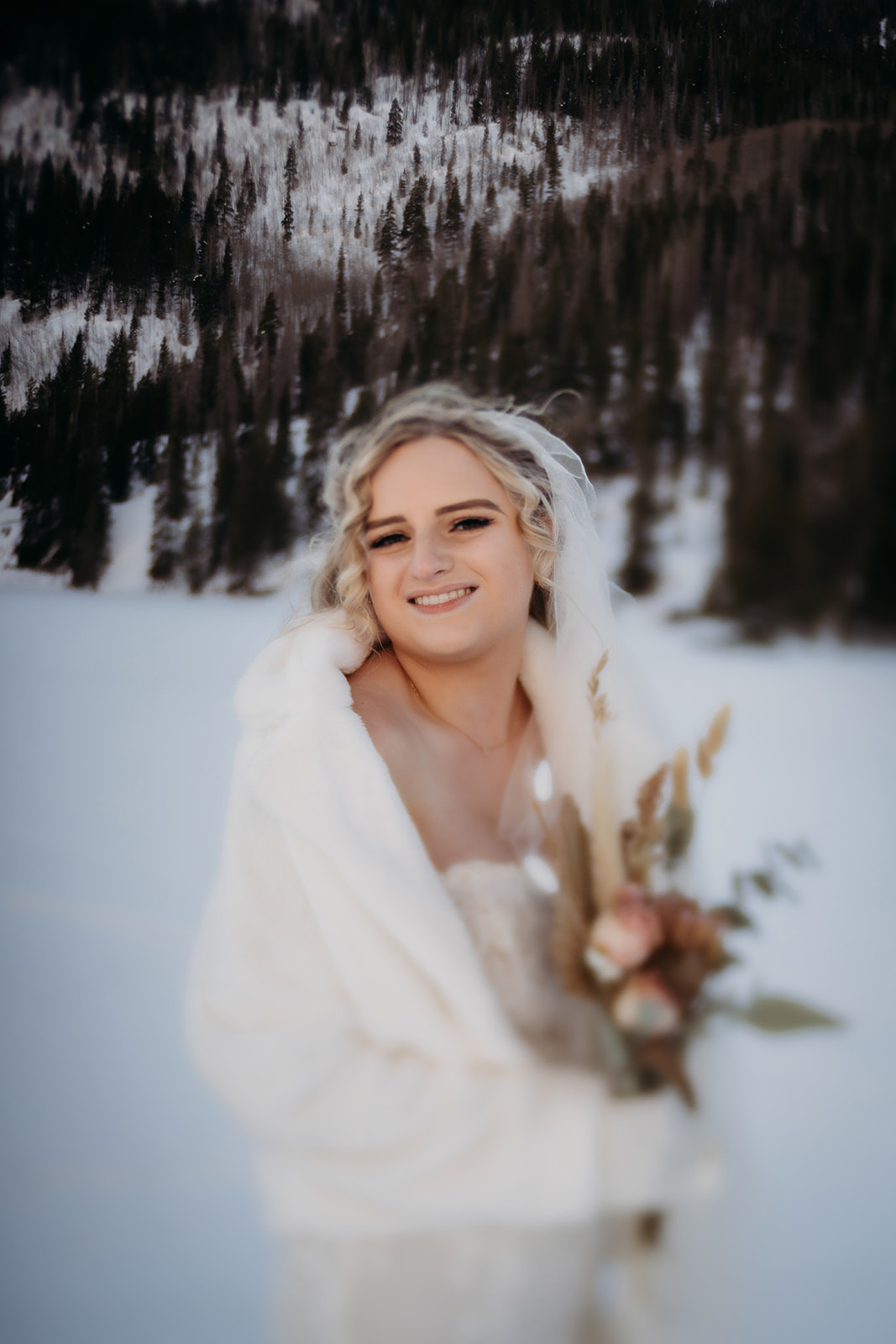 Bride smiling in a soft white faux fur jacket, holding a rustic bouquet in the snowy woods—perfect inspiration for winter wedding outfits.