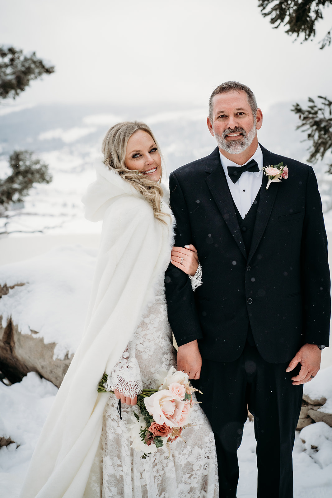 Bride in a lace gown and dramatic white cape standing with her groom in a snowy mountaintop setting.