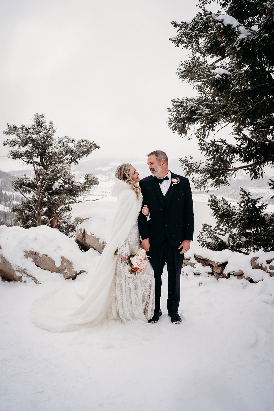 Bride in a detailed lace gown with a flowing cape walking arm in arm with her groom, surrounded by a winter wonderland.