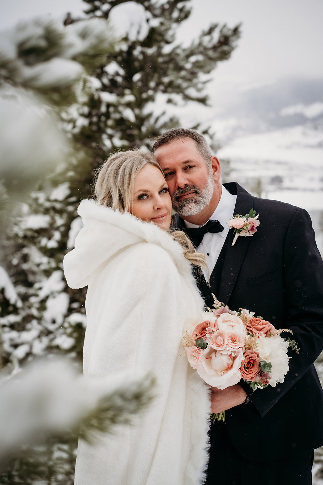 Bride in a white hooded cape and groom in a black tuxedo snuggling in a snowy forest, capturing the elegance of winter wedding outfits.