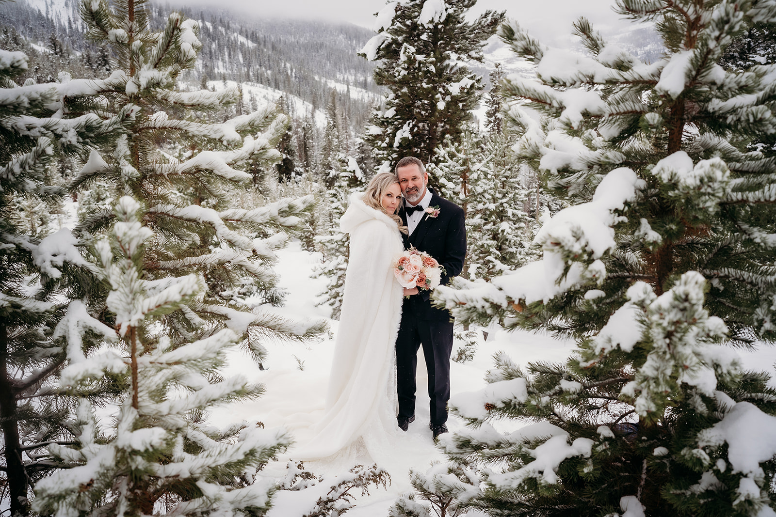 Bride and groom snuggled together in a snowy forest, showcasing winter wedding outfits perfect for a cozy mountain ceremony.