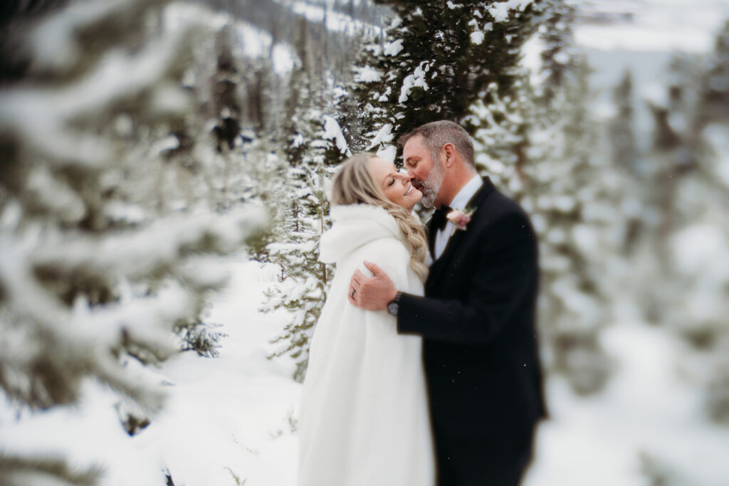 Groom whispering sweetly to his bride, who is wrapped in a white winter cape, surrounded by snow-covered trees—an intimate moment showcasing romantic winter wedding outfits.