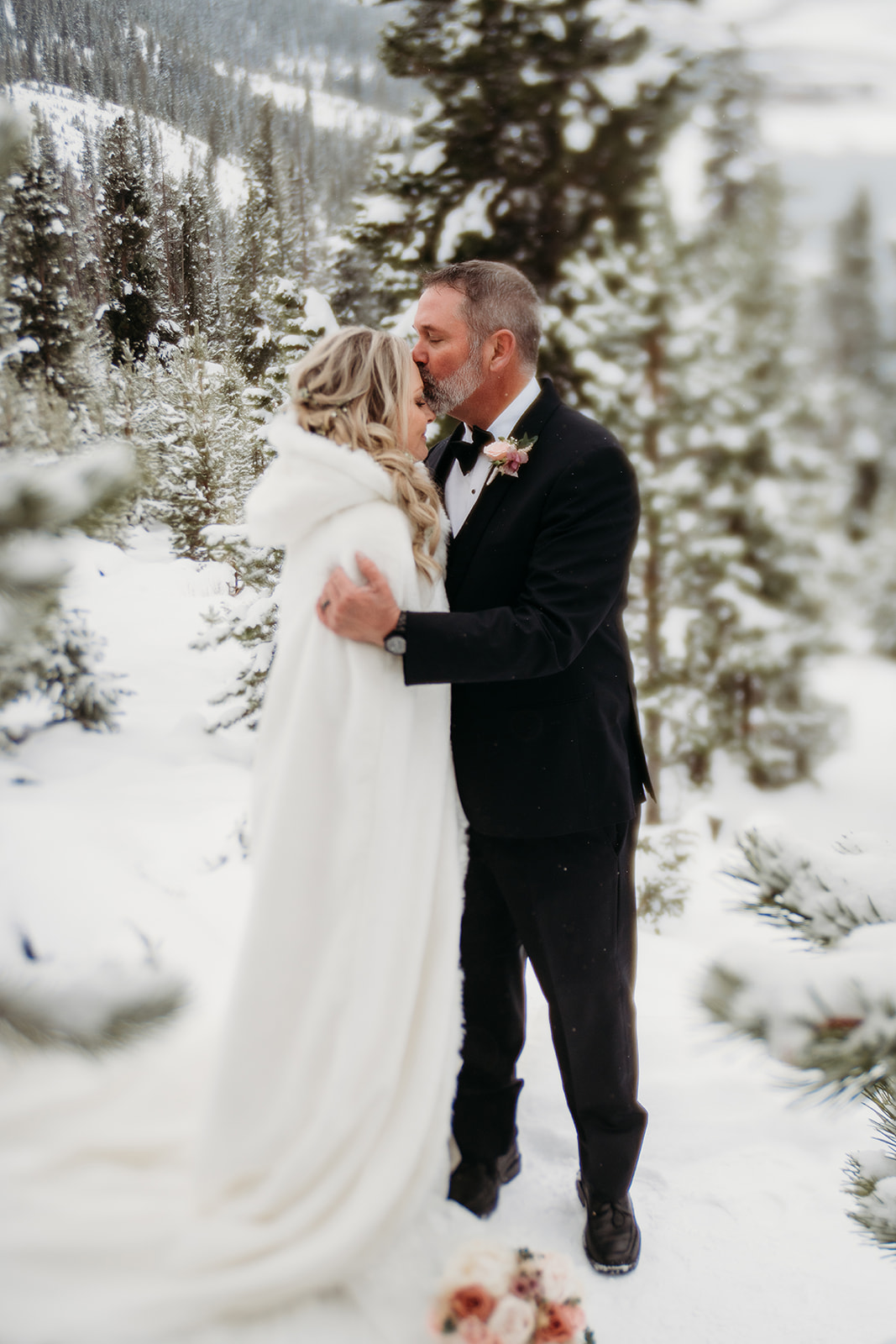 Groom in tuxedo leaning in toward bride wrapped in a hooded white cape, surrounded by Colorado’s snowy pine trees.