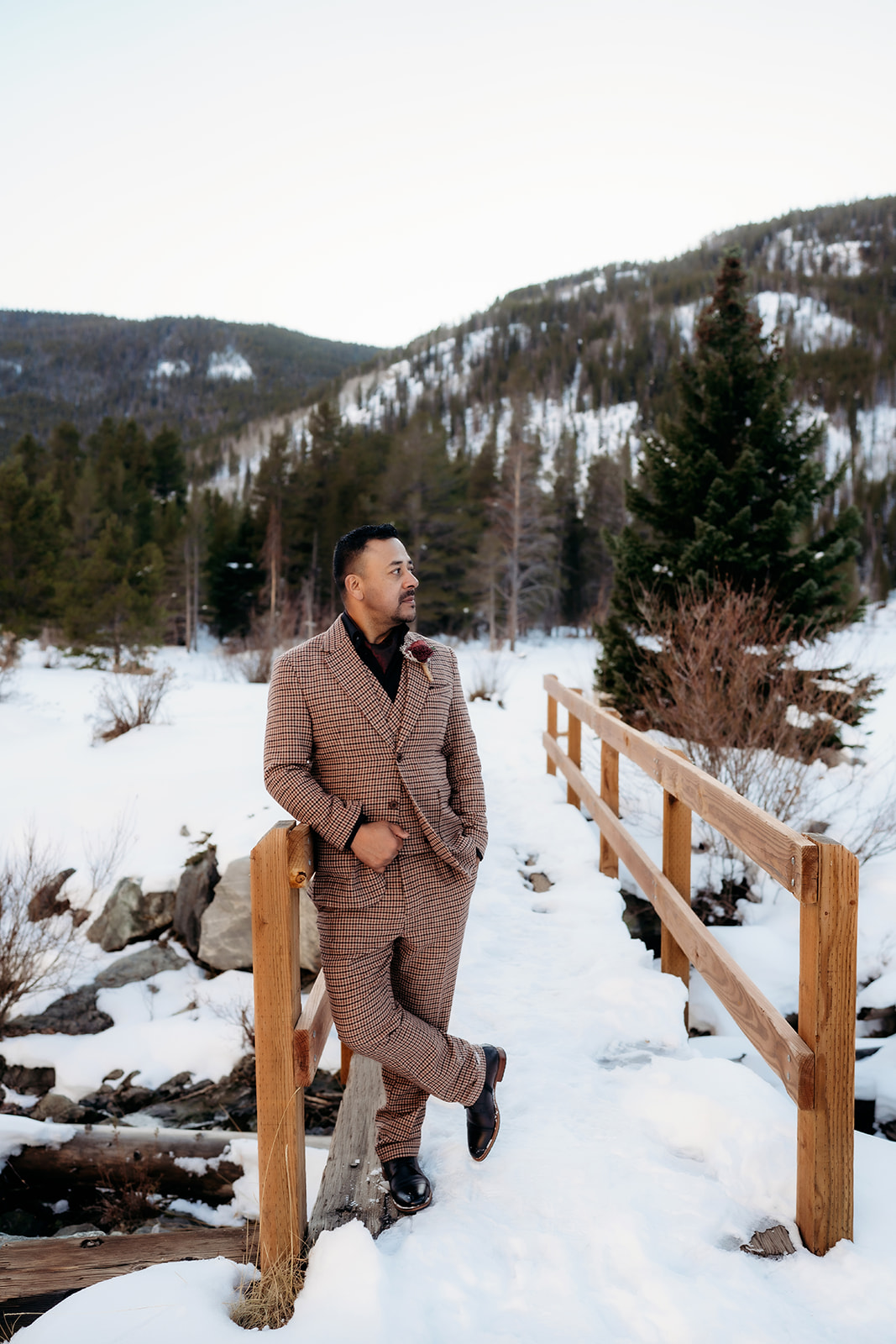 Groom beaming on a rustic wooden bridge, his modern brown plaid suit standing out beautifully in a snowy mountain scene.