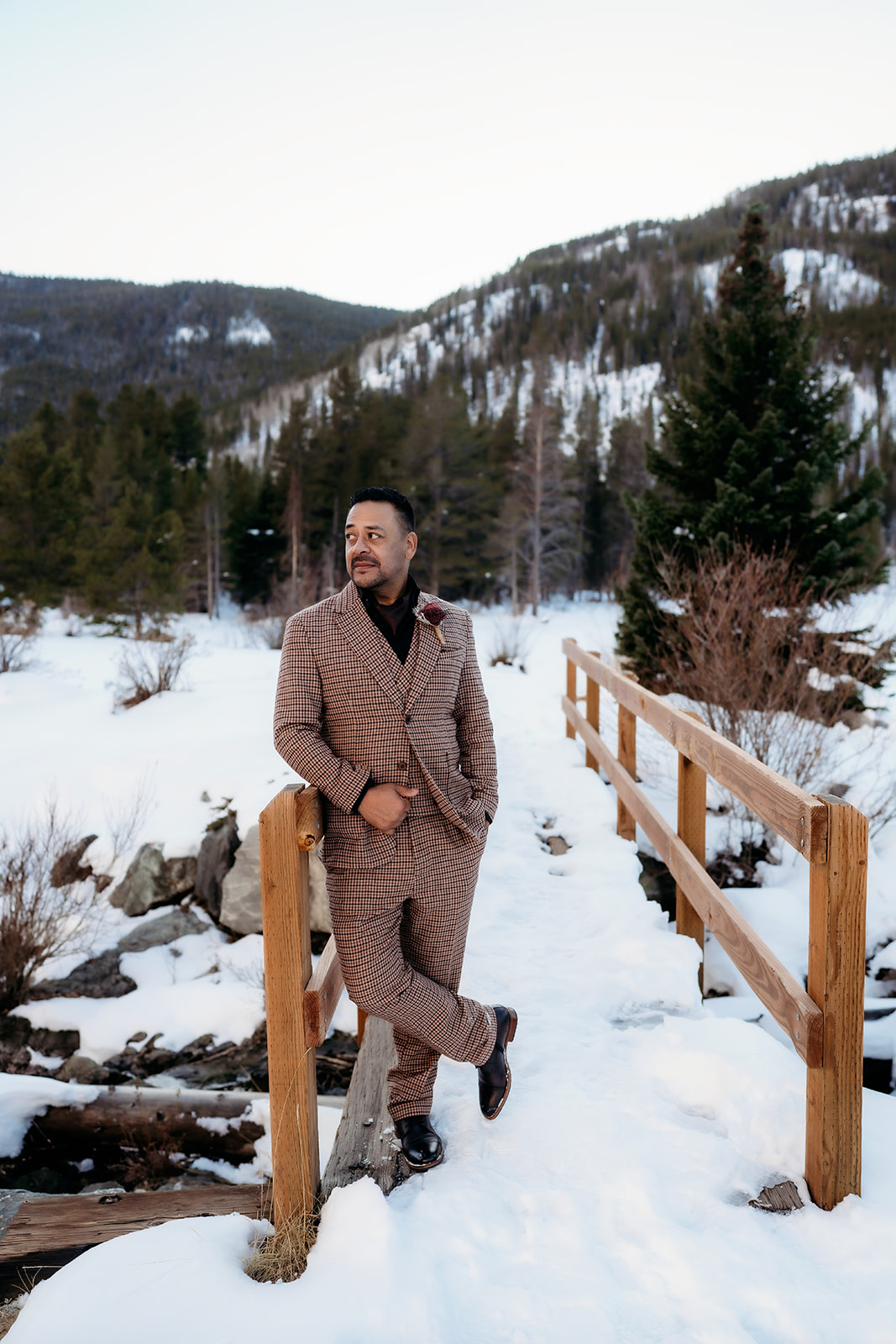 Groom in a stylish brown plaid suit standing on a wooden bridge with a snow-covered Colorado mountain backdrop, a modern take on winter wedding outfits.