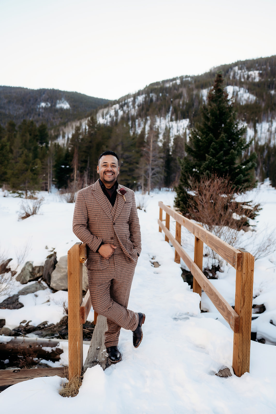 Groom in a brown plaid suit posing on a wooden bridge with a snow-dusted mountain landscape behind him.