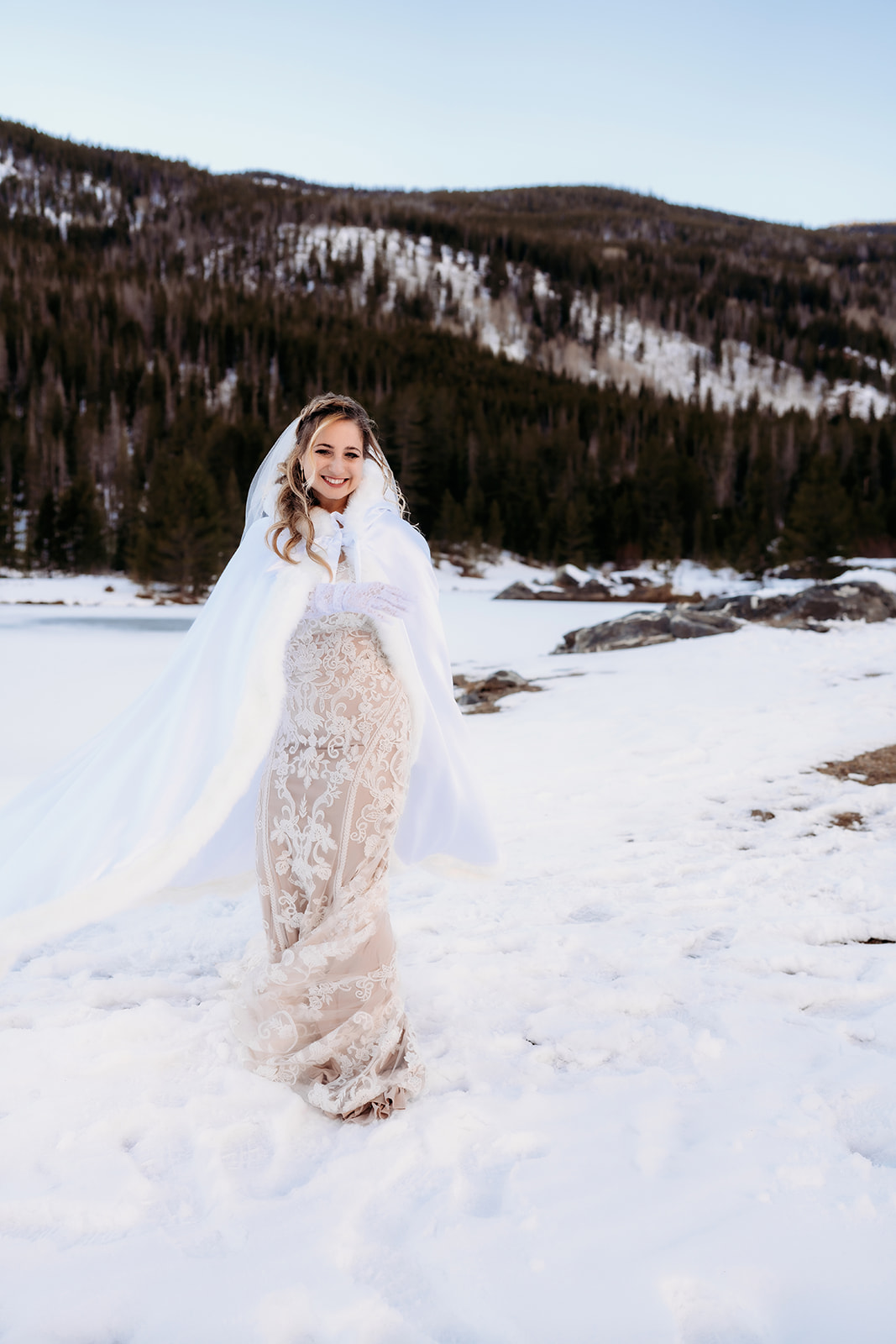 Bride in a sheer embroidered gown with a flowing white cape, standing on a snowy lakeside trail—a showstopping winter wedding outfit.