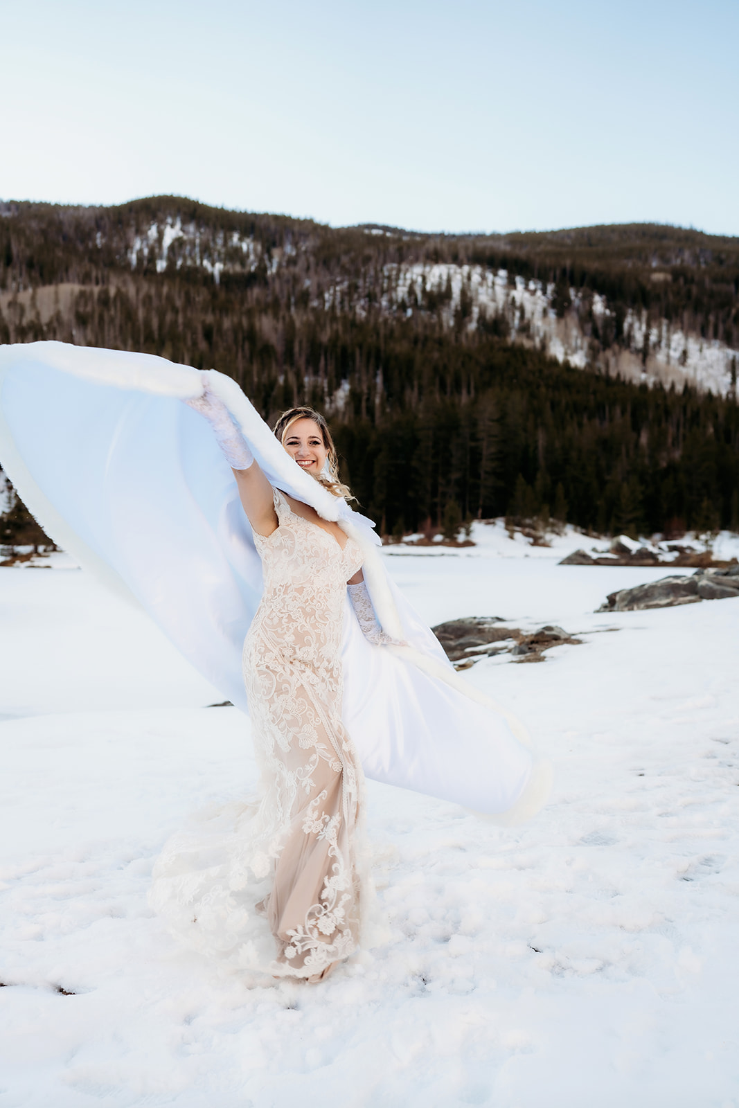 Joyful bride twirls in the snow wearing a fitted lace gown and dramatic white cape, capturing the essence of playful winter wedding outfits.