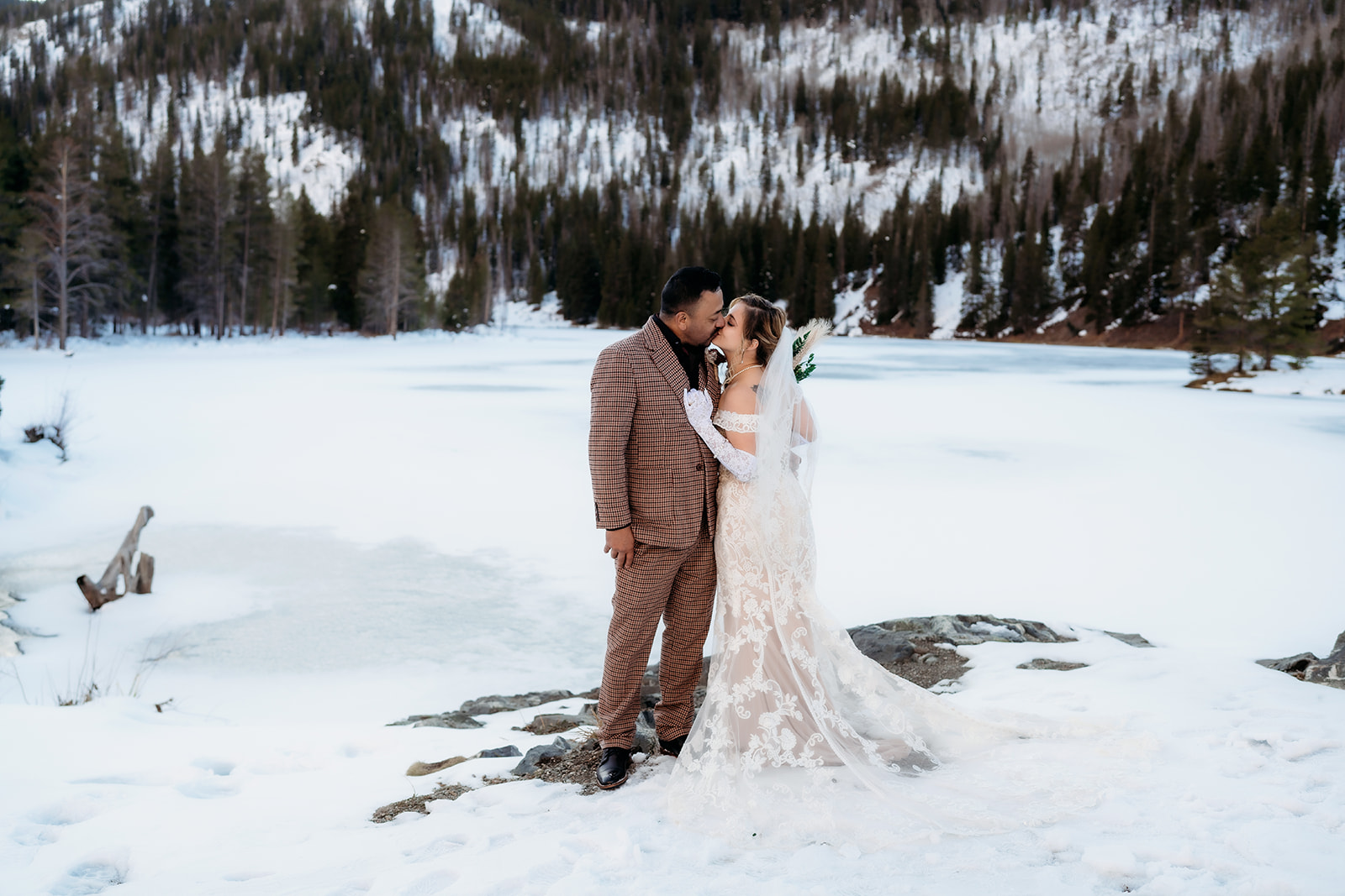 Bride in an off-shoulder lace gown and groom in a plaid suit share a kiss beside a snow-covered lake, dressed in stylish winter wedding outfits.